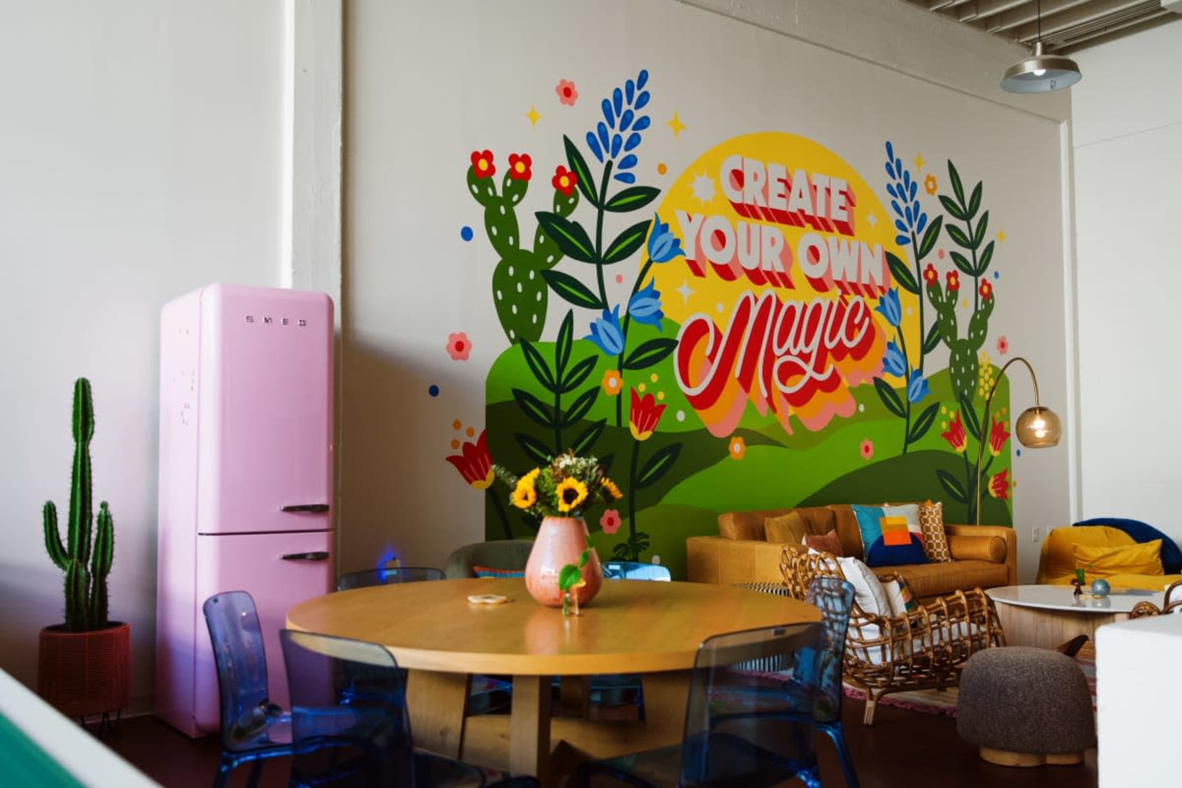 The image shows a vibrant cafe interior with a colorful mural, a pink refrigerator, and a mix of seating options, including a round dining table and a cozy couch.