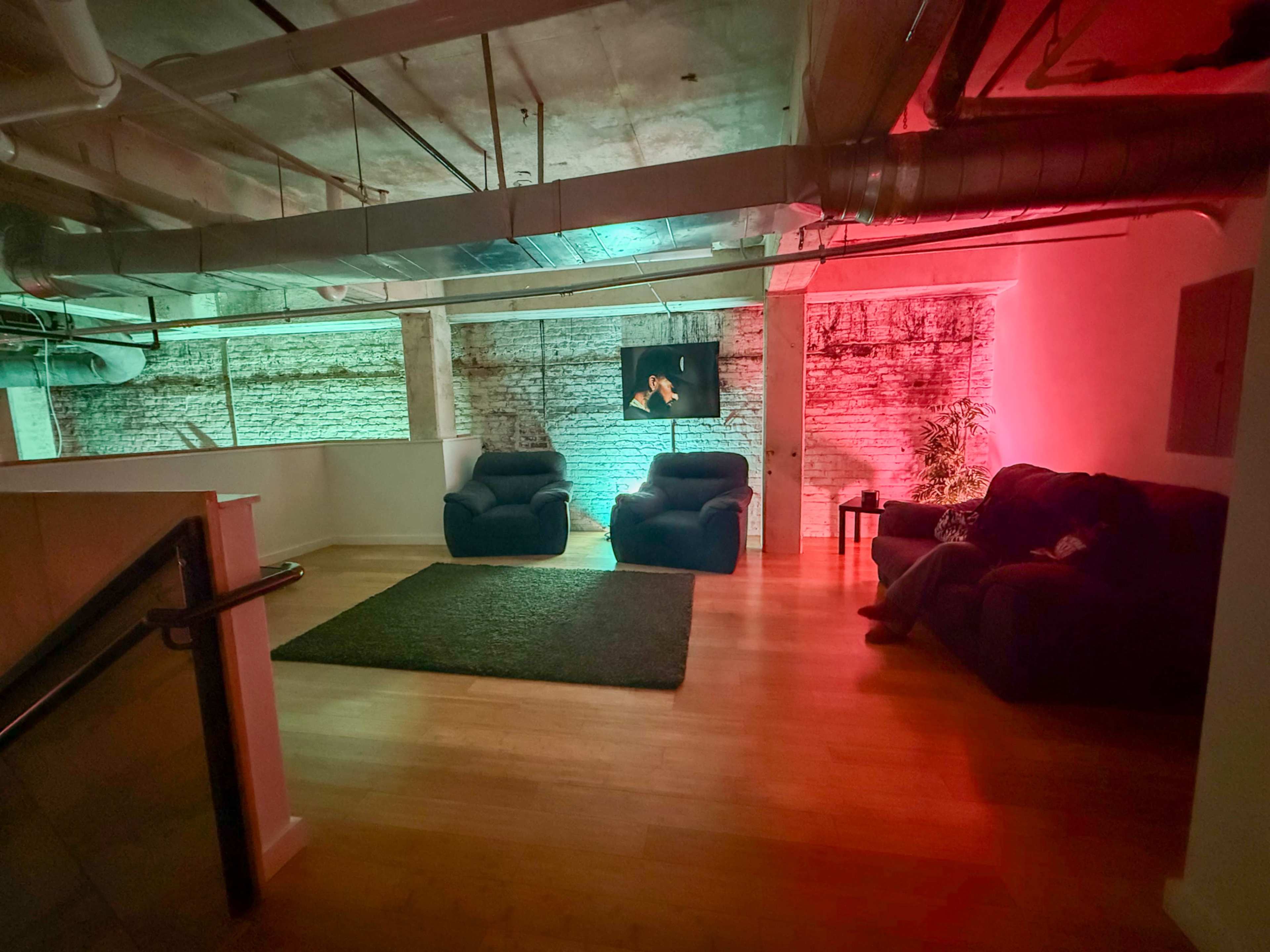 The image shows a spacious indoor room with two dark sofas, a large area rug, and ambient lighting in green and red hues.