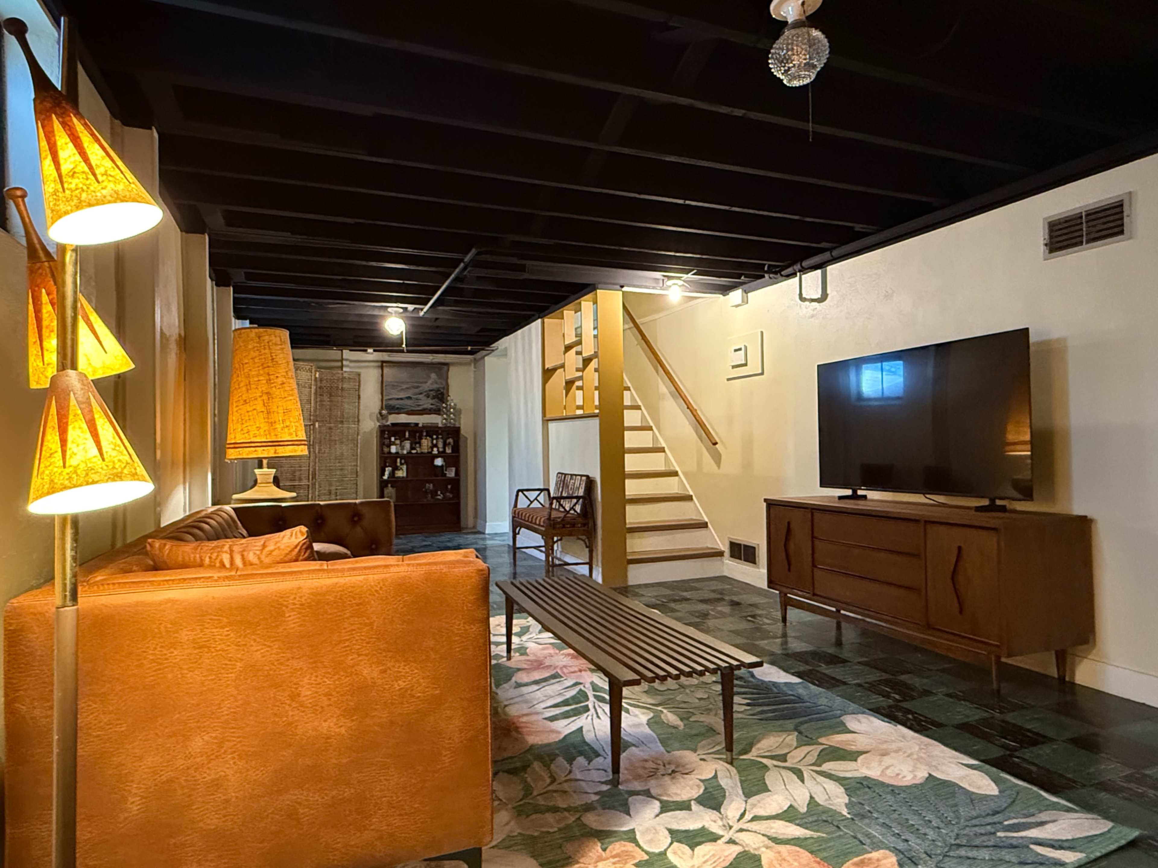 The image depicts a cozy basement living area featuring a brown sofa, a wooden coffee table, a TV stand, and decorative lamps, with a staircase visible in the background.
