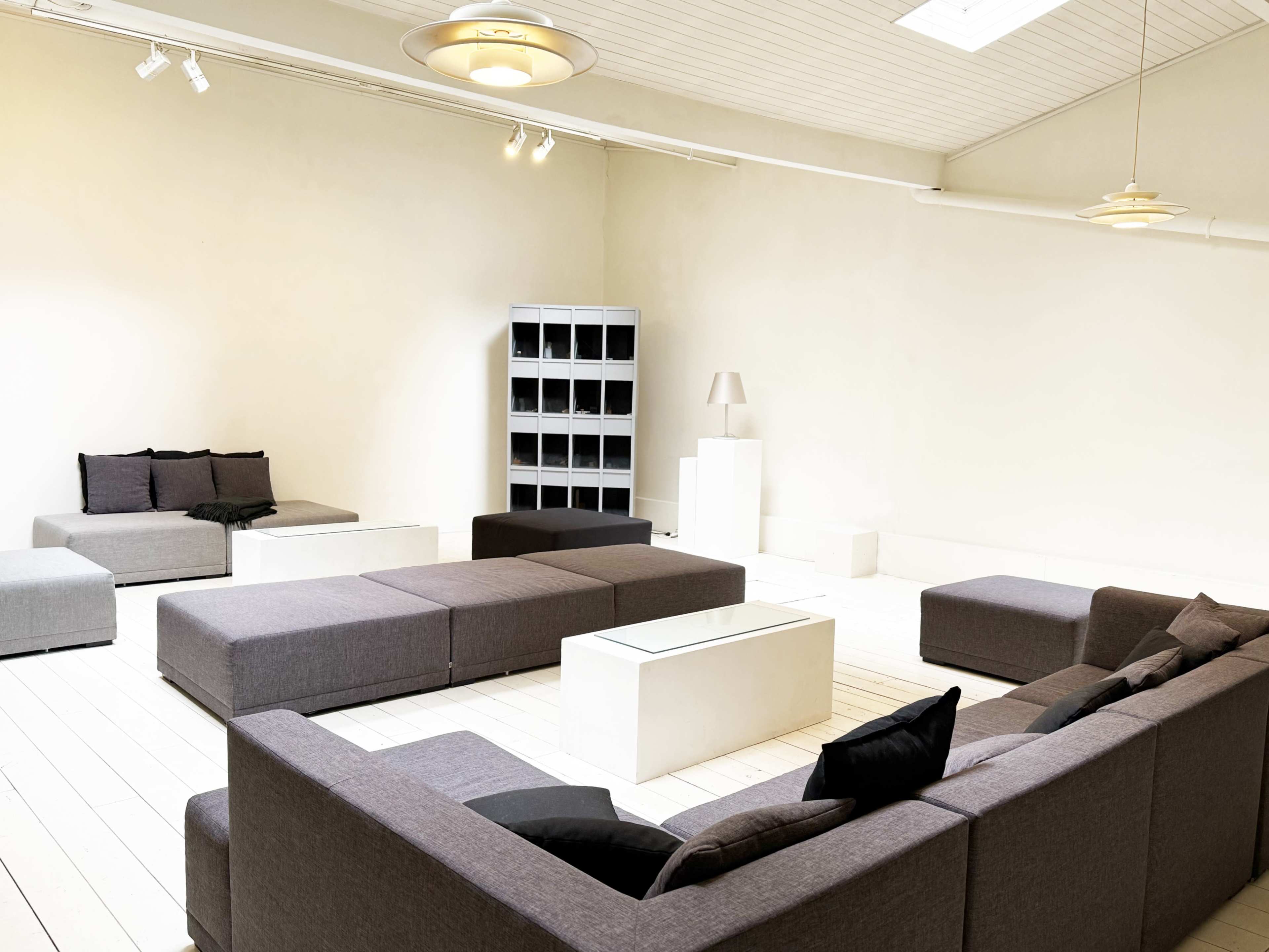 The image shows a modern, minimalist living space featuring several gray modular sofas arranged around a central glass coffee table with a light-colored bookshelf and lamp in the background.