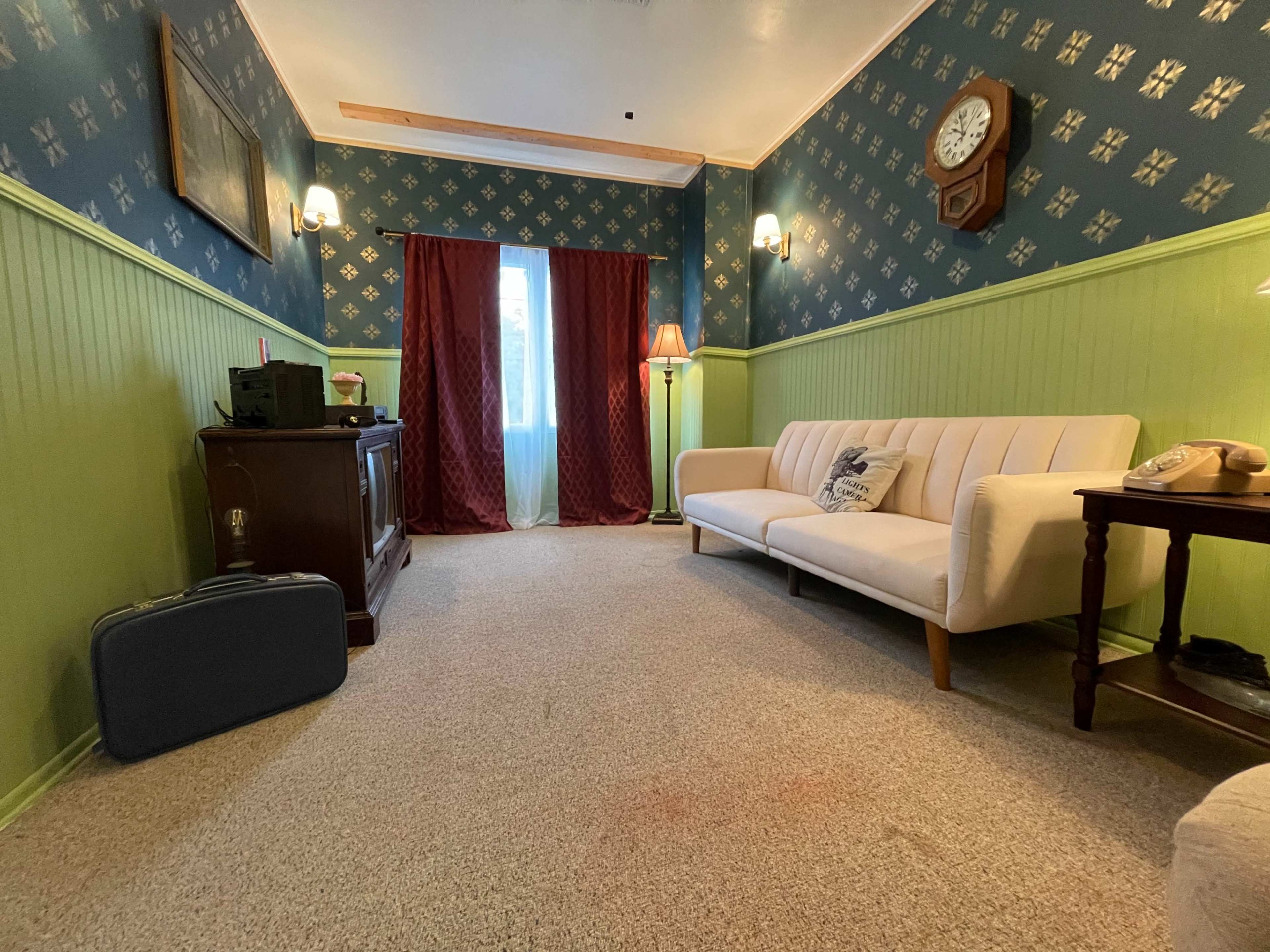The room features a beige sofa, a wooden side table, a vintage clock on the wall, and a window with red curtains, all surrounded by patterned blue wallpaper and green wainscoting.
