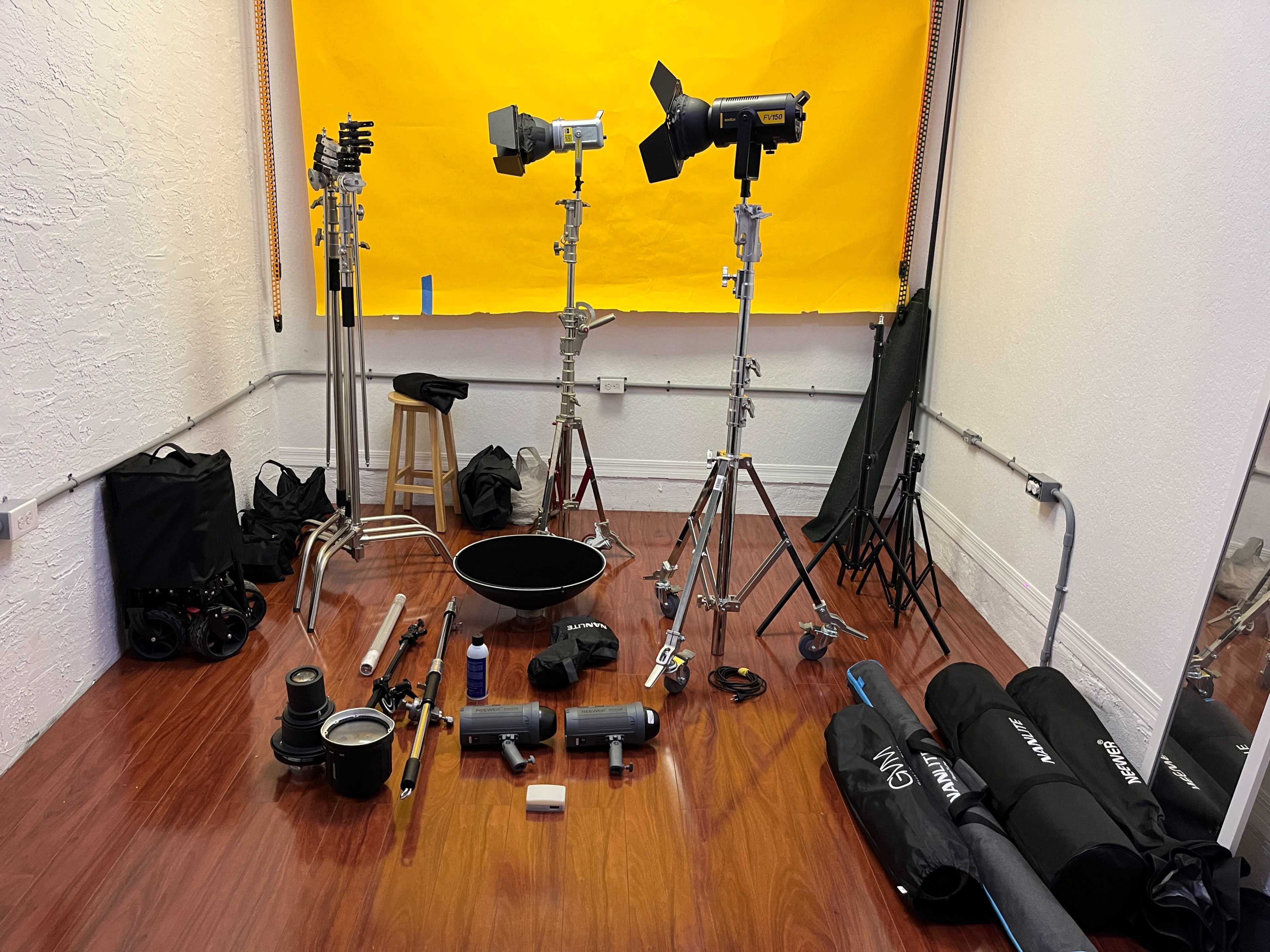 A photography studio with various lighting equipment, stands, and bags arranged neatly on a wooden floor in front of a bright yellow backdrop.