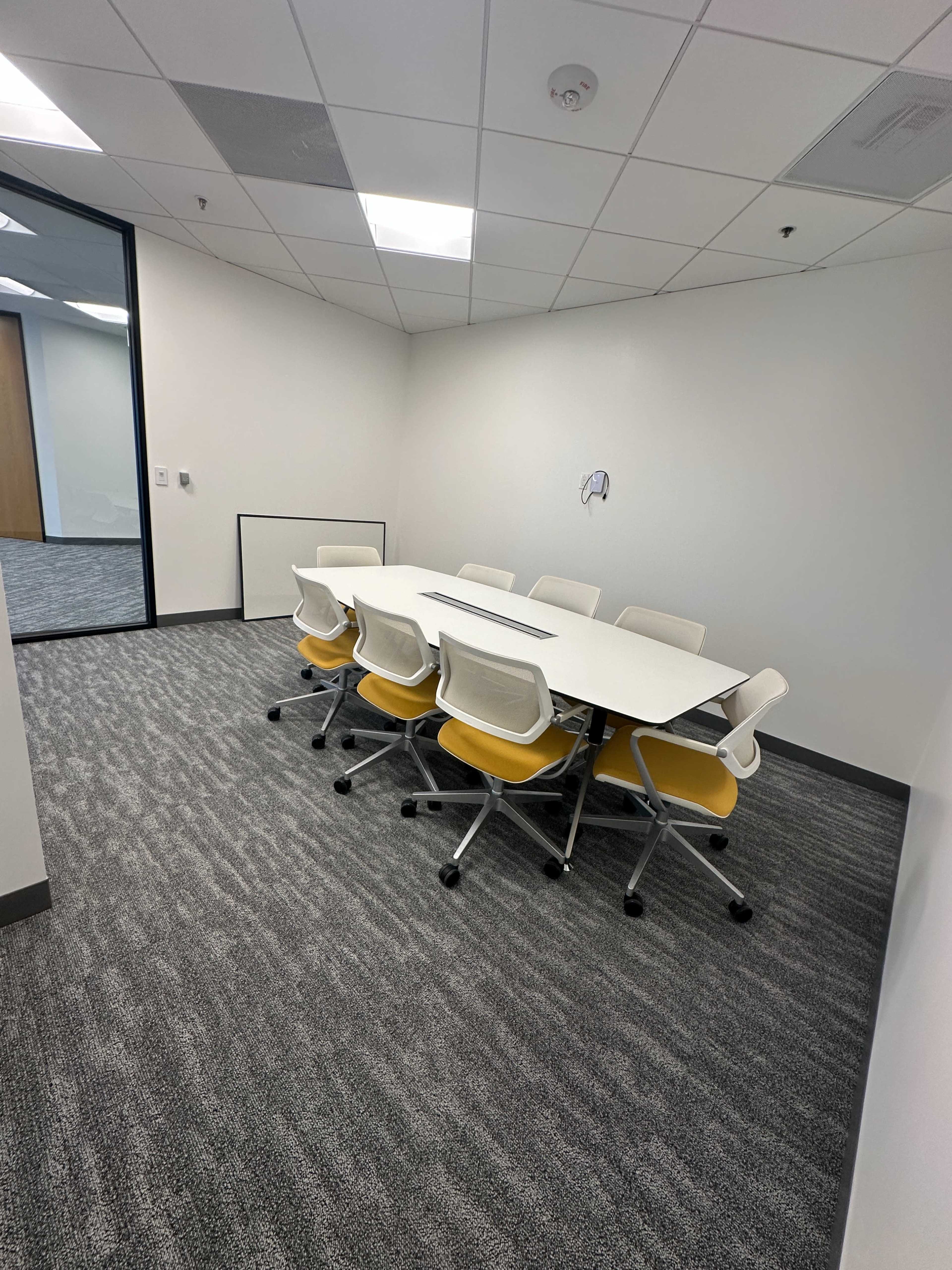 The image shows a modern conference room with a large white table surrounded by yellow and white chairs, and gray carpet flooring.