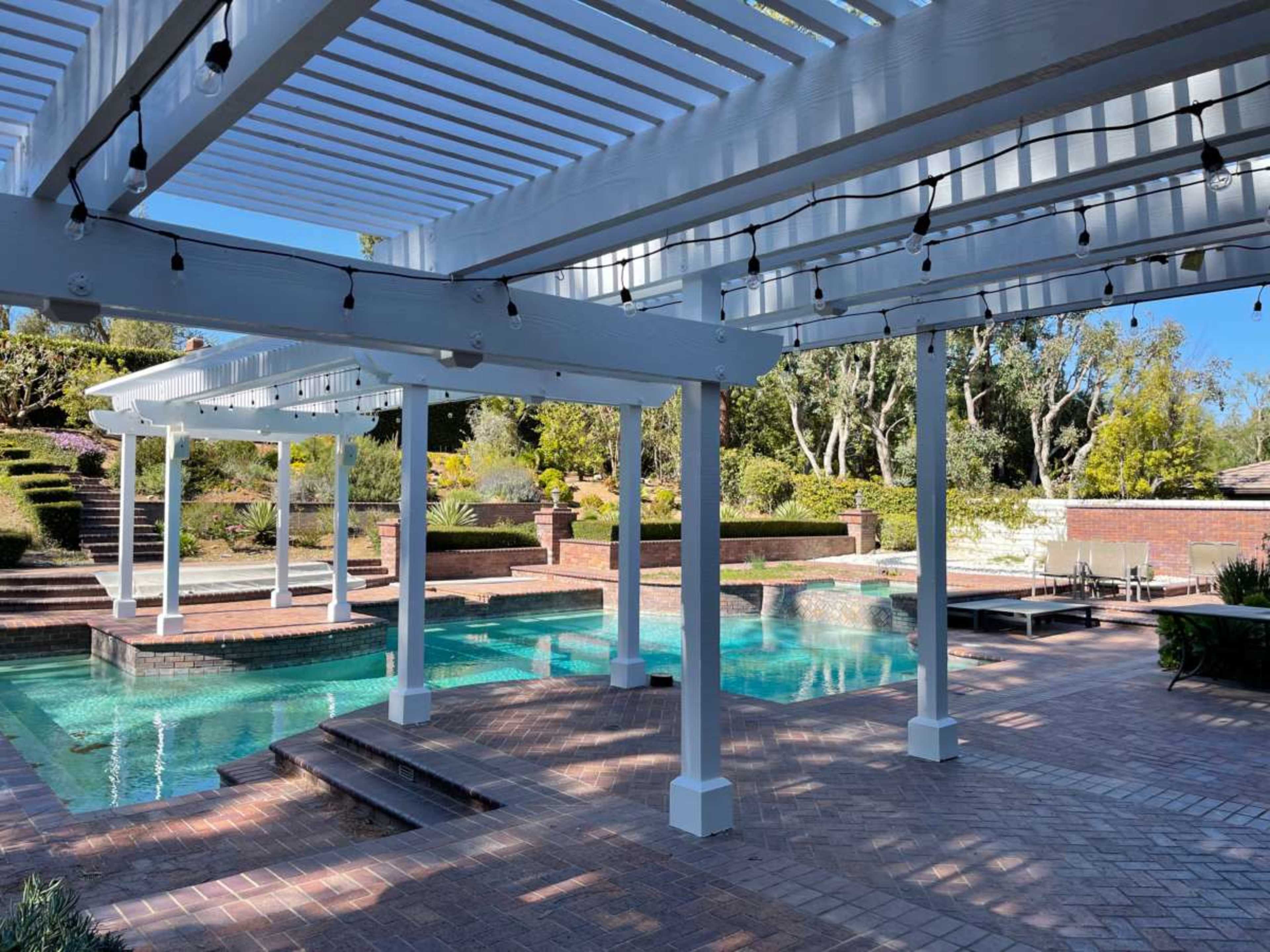 The image shows a backyard pool area with a covered deck structure, surrounded by landscaped gardens and pathways.
