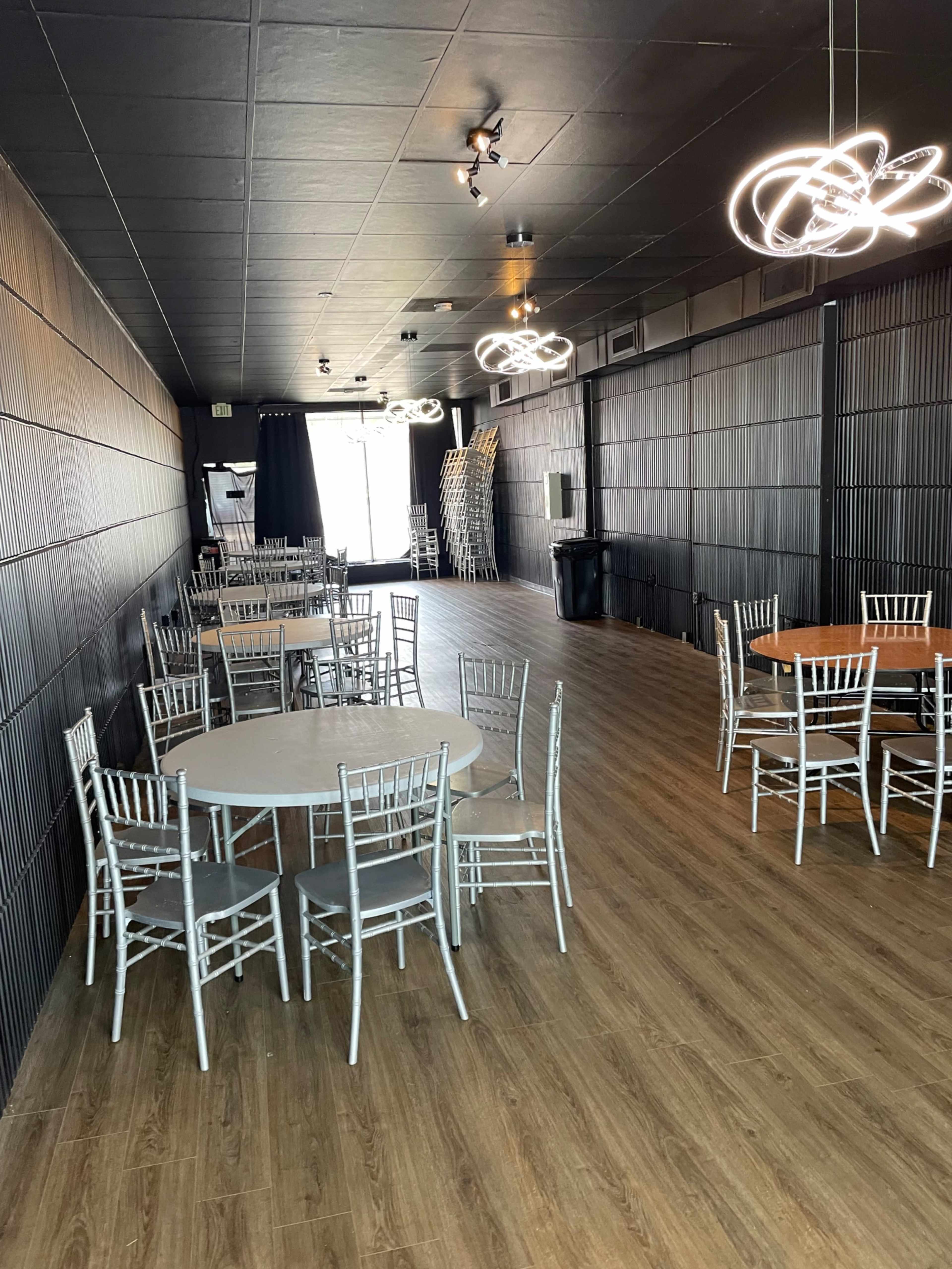 A spacious indoor venue with several round tables and chairs arranged on a wooden floor, illuminated by contemporary light fixtures.
