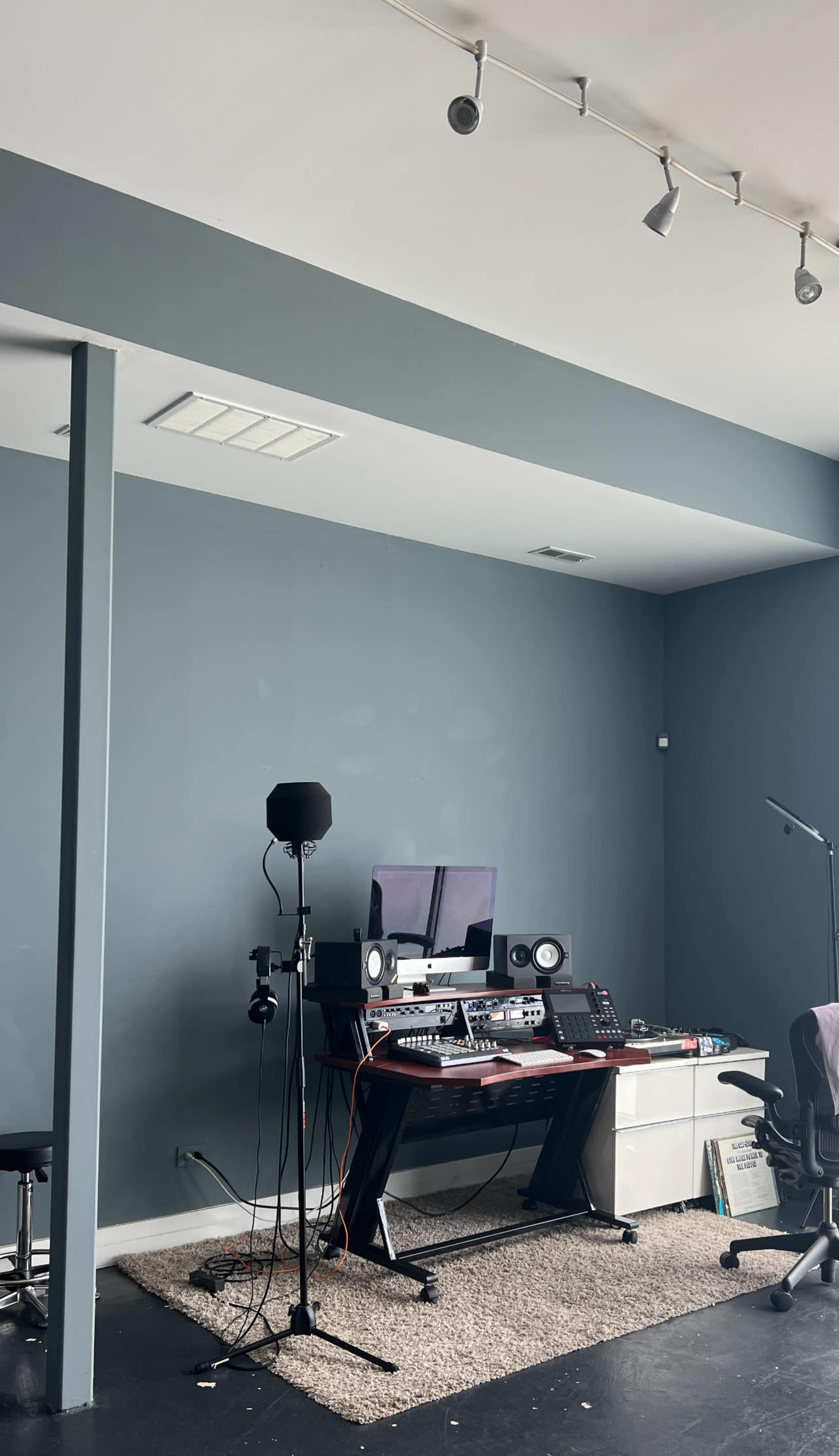 A music production studio features a desk with audio equipment, monitors, and a microphone setup against a blue wall.