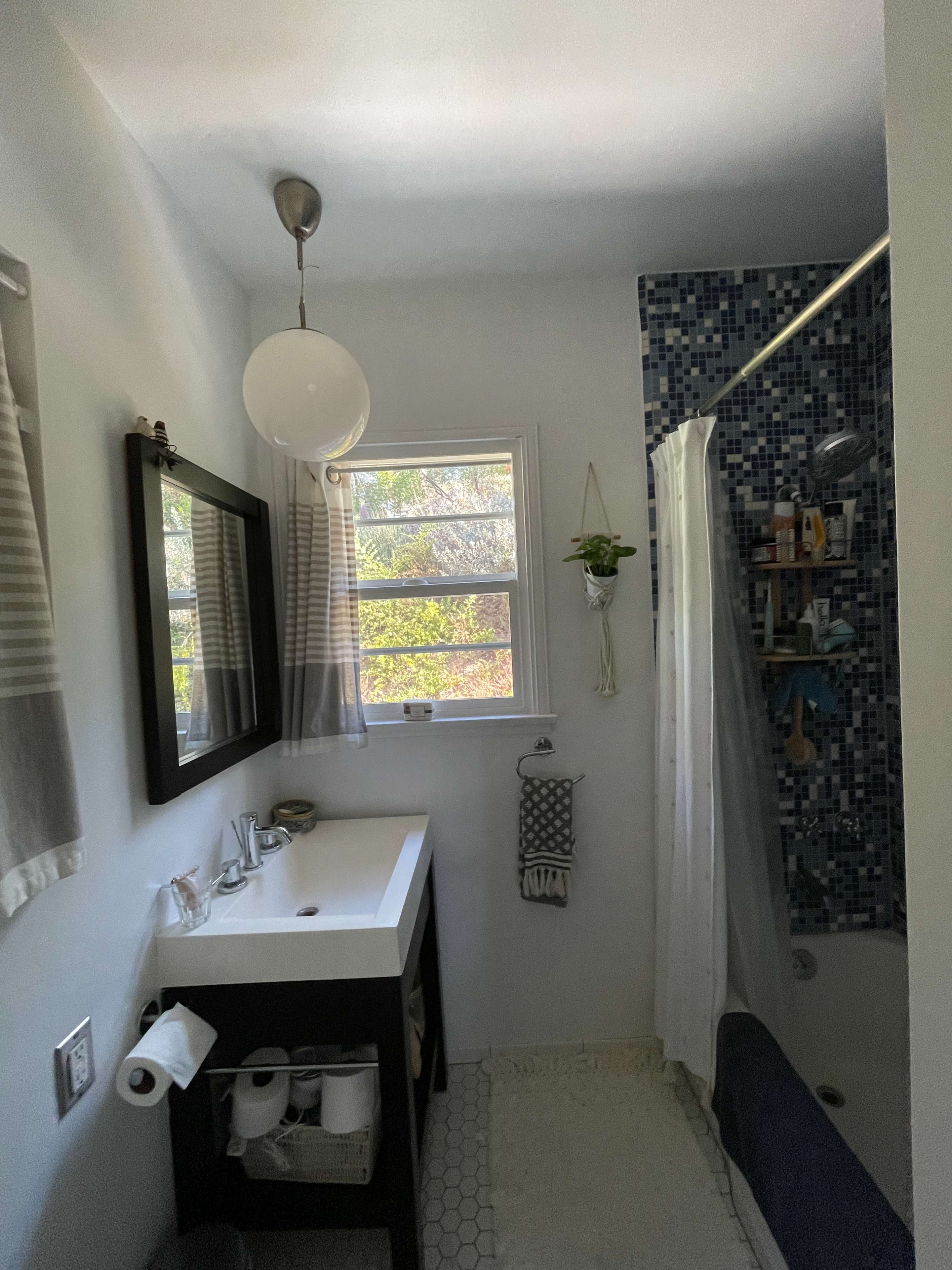A small bathroom featuring a white vanity with a mirror, a shower area with tiled walls, and a window that provides natural light.