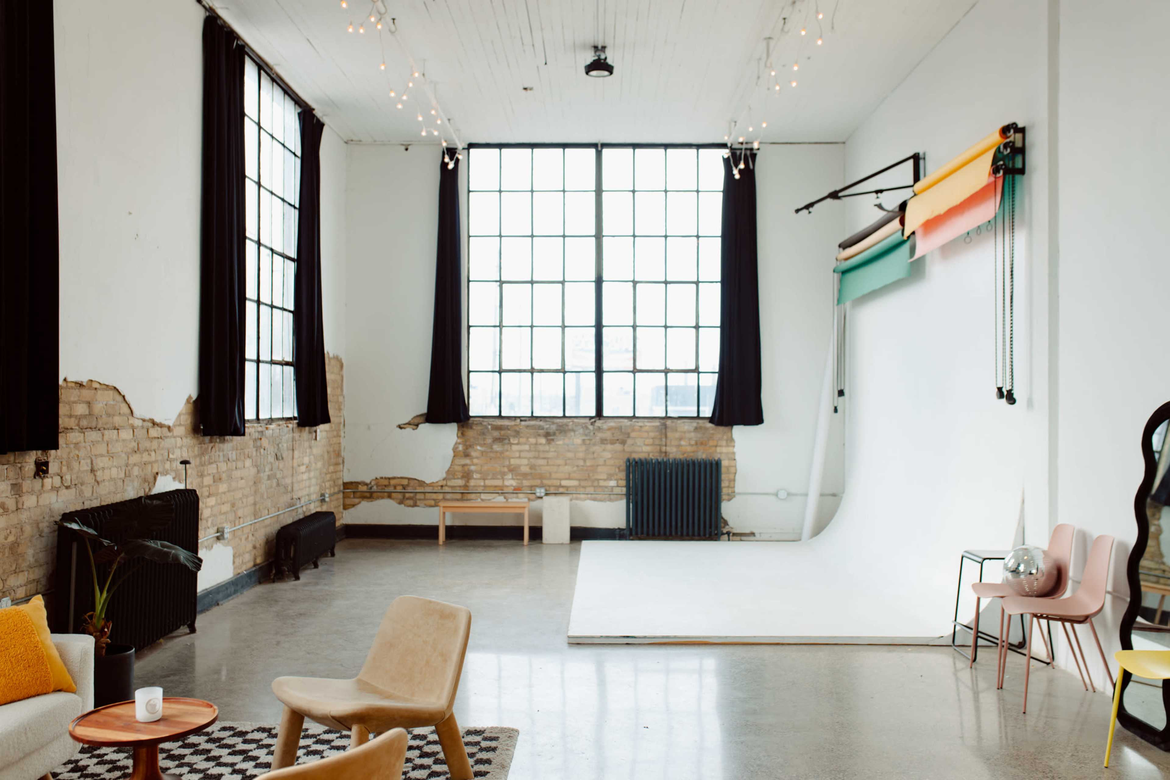 The image shows a spacious, well-lit room with large windows, exposed brick walls, and a simple backdrop setup for photography.