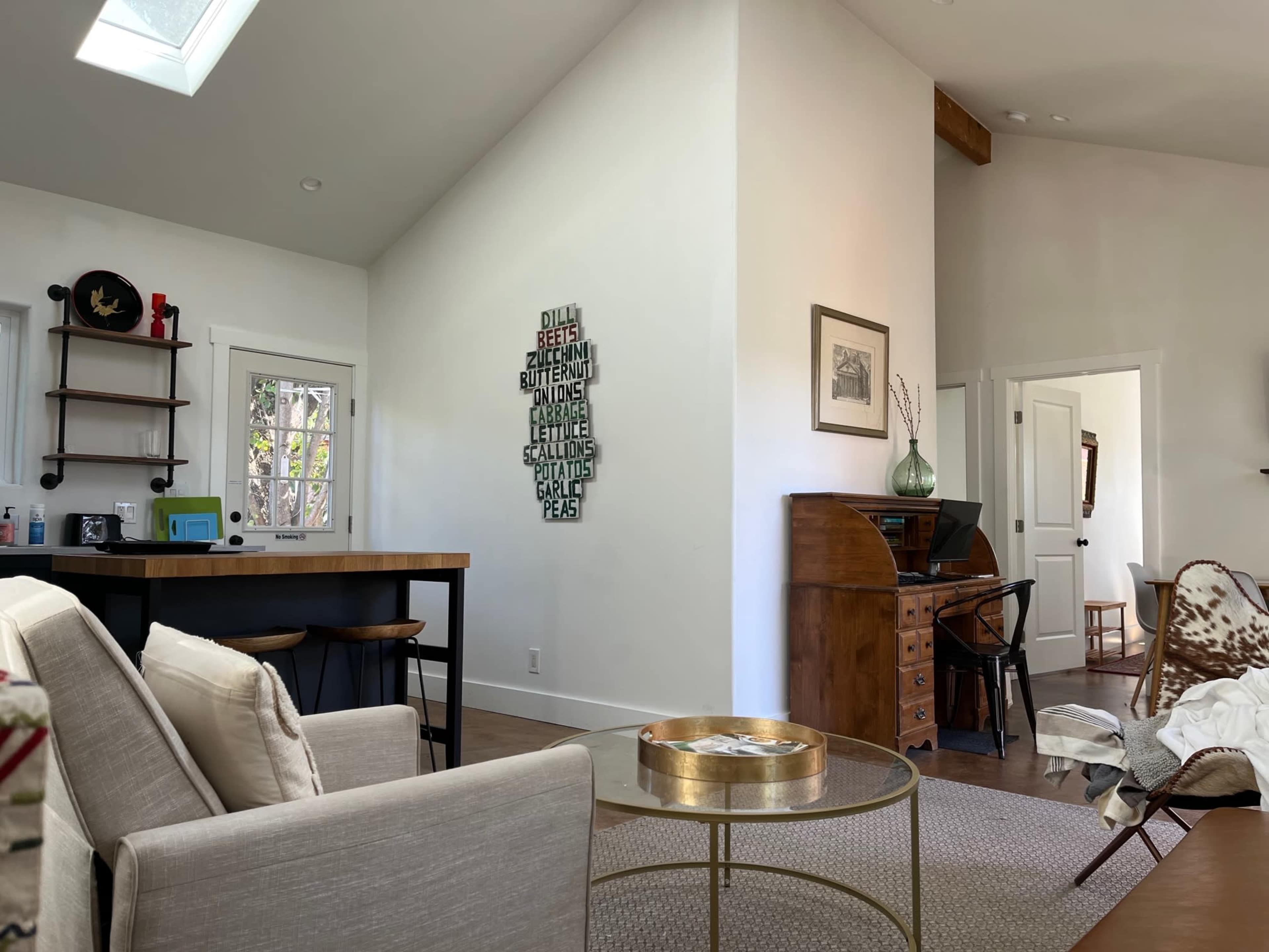 A modern interior space featuring a living area with a sofa, a dining table with stools, and a desk against the wall, adorned with decorative items and a wall-mounted art piece made of metal letters.
