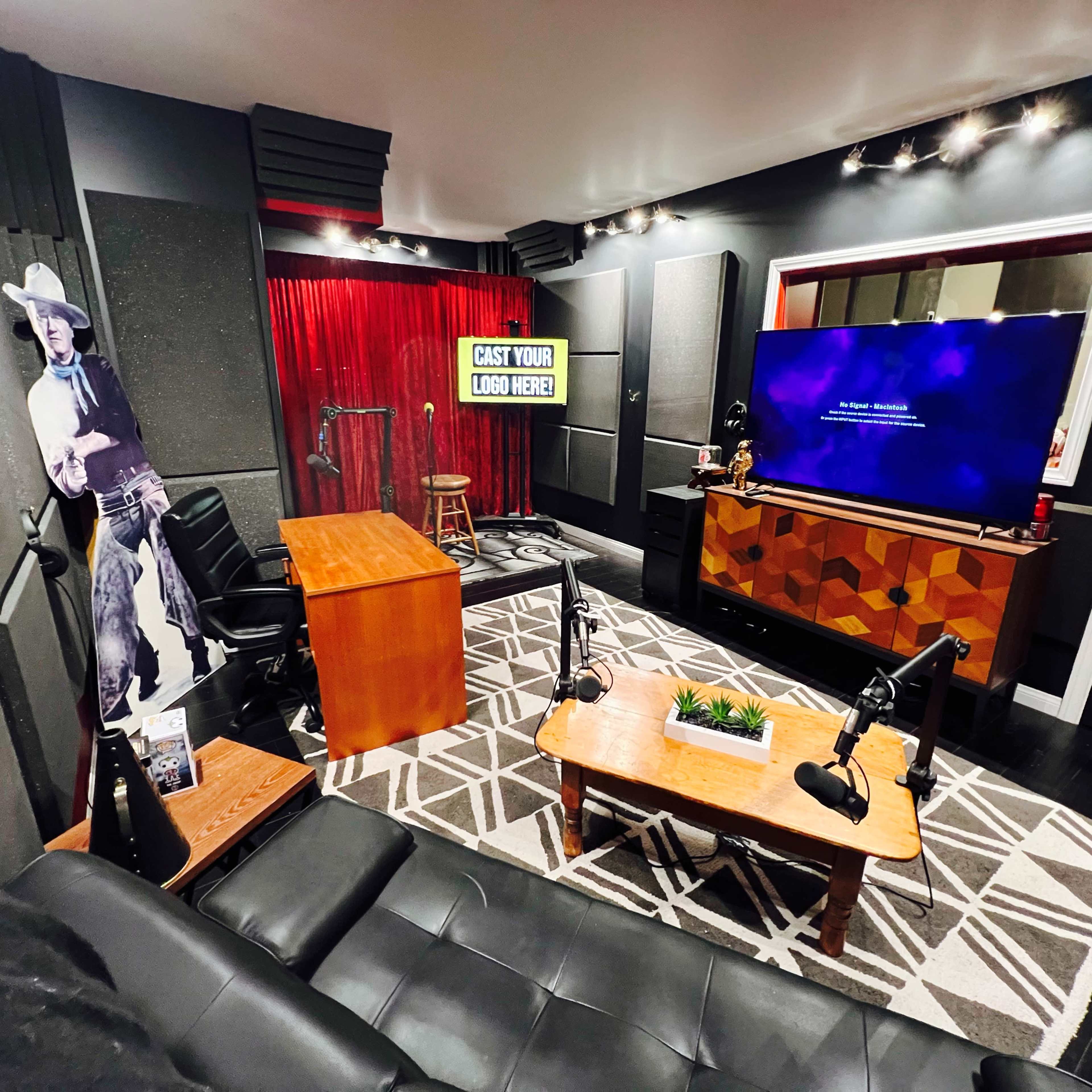 The image features a recording studio with black walls, a wooden desk, two chairs, a large television, and a cowboy cutout in the corner.