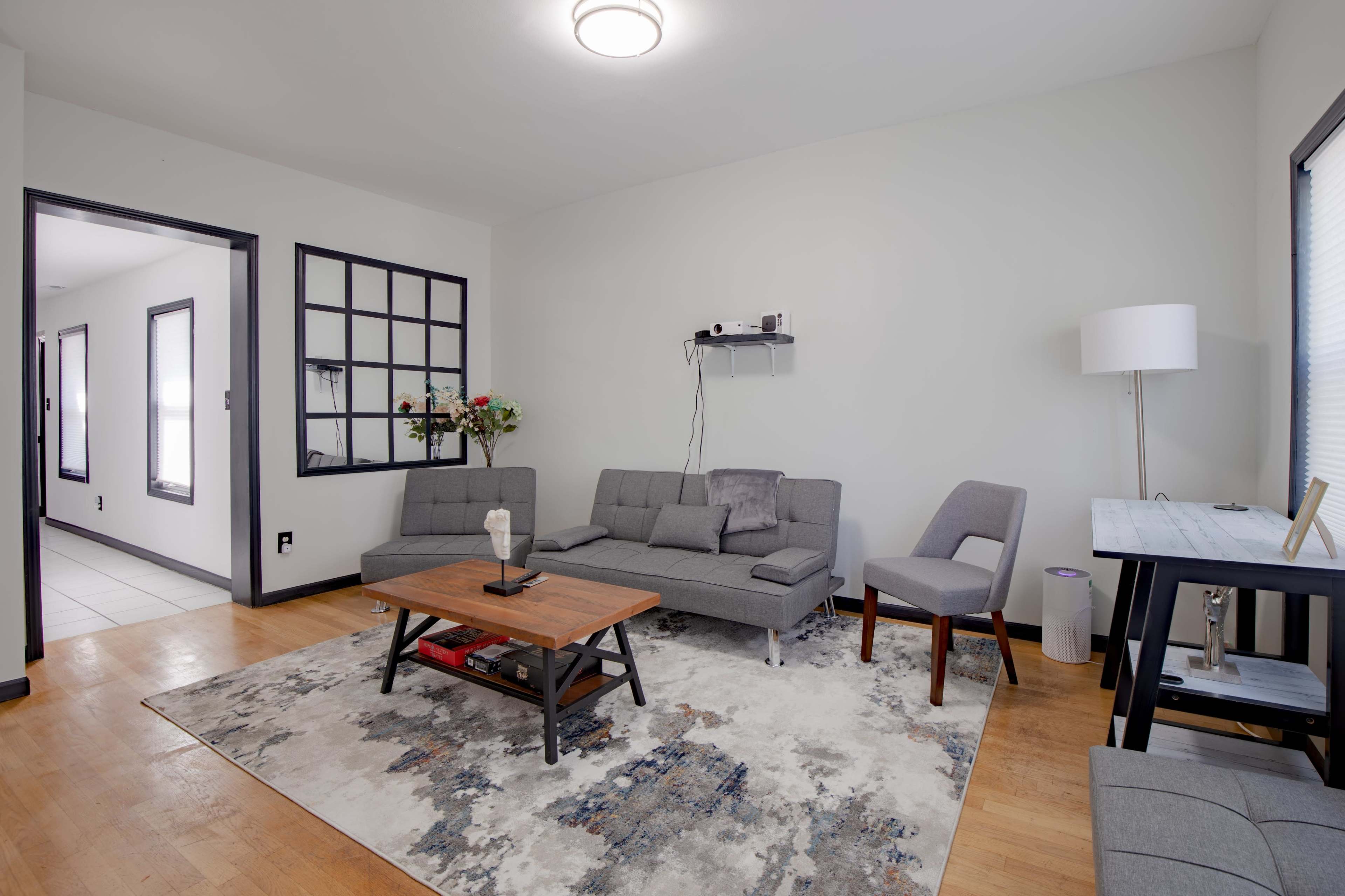 A modern living room features a gray sectional sofa, a wooden coffee table, and a side chair, with a large area rug on the wooden floor and a lamp next to a small table.