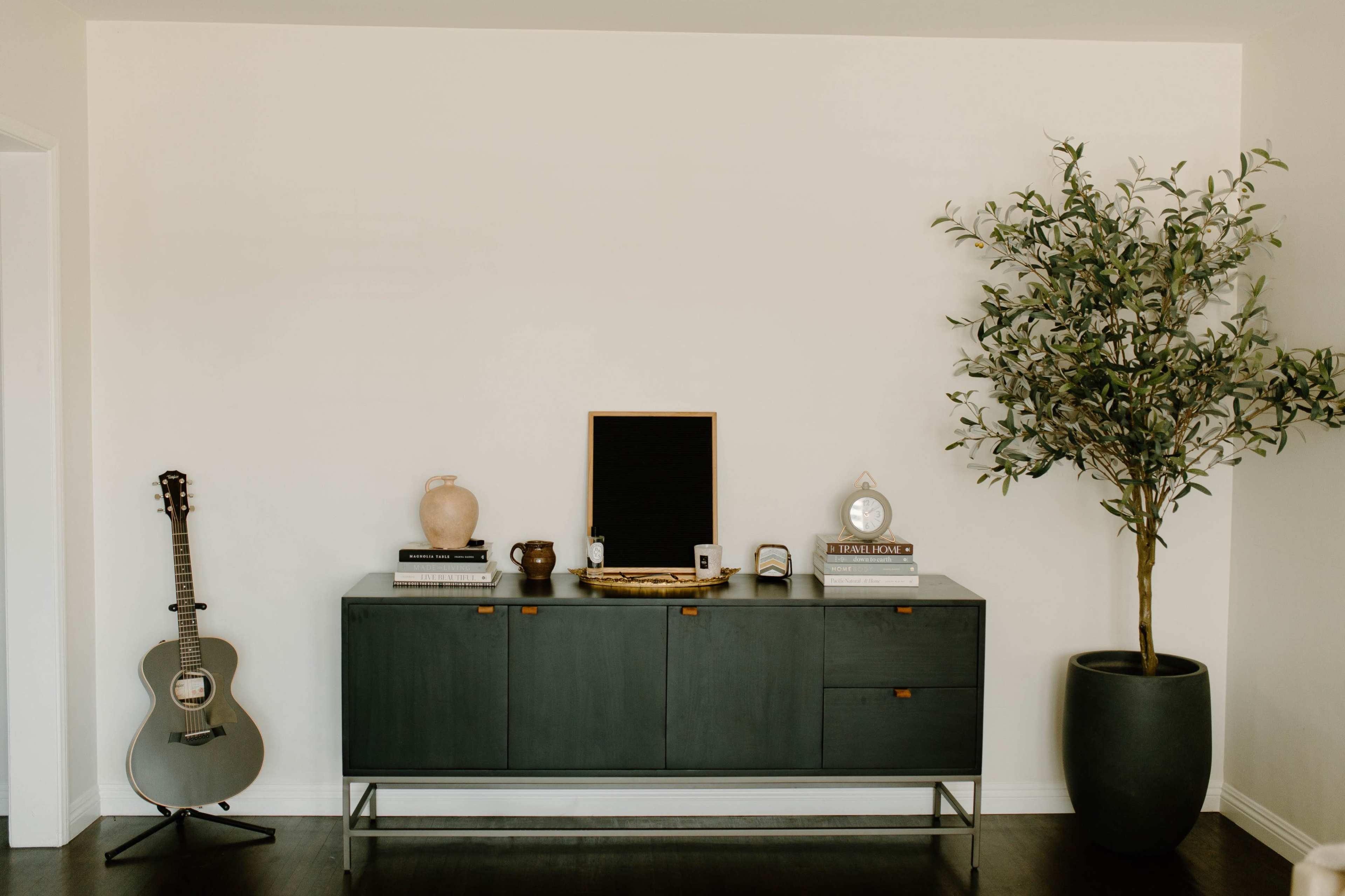 A dark sideboard with decorative items, including a guitar and a potted tree, is positioned against a plain wall.