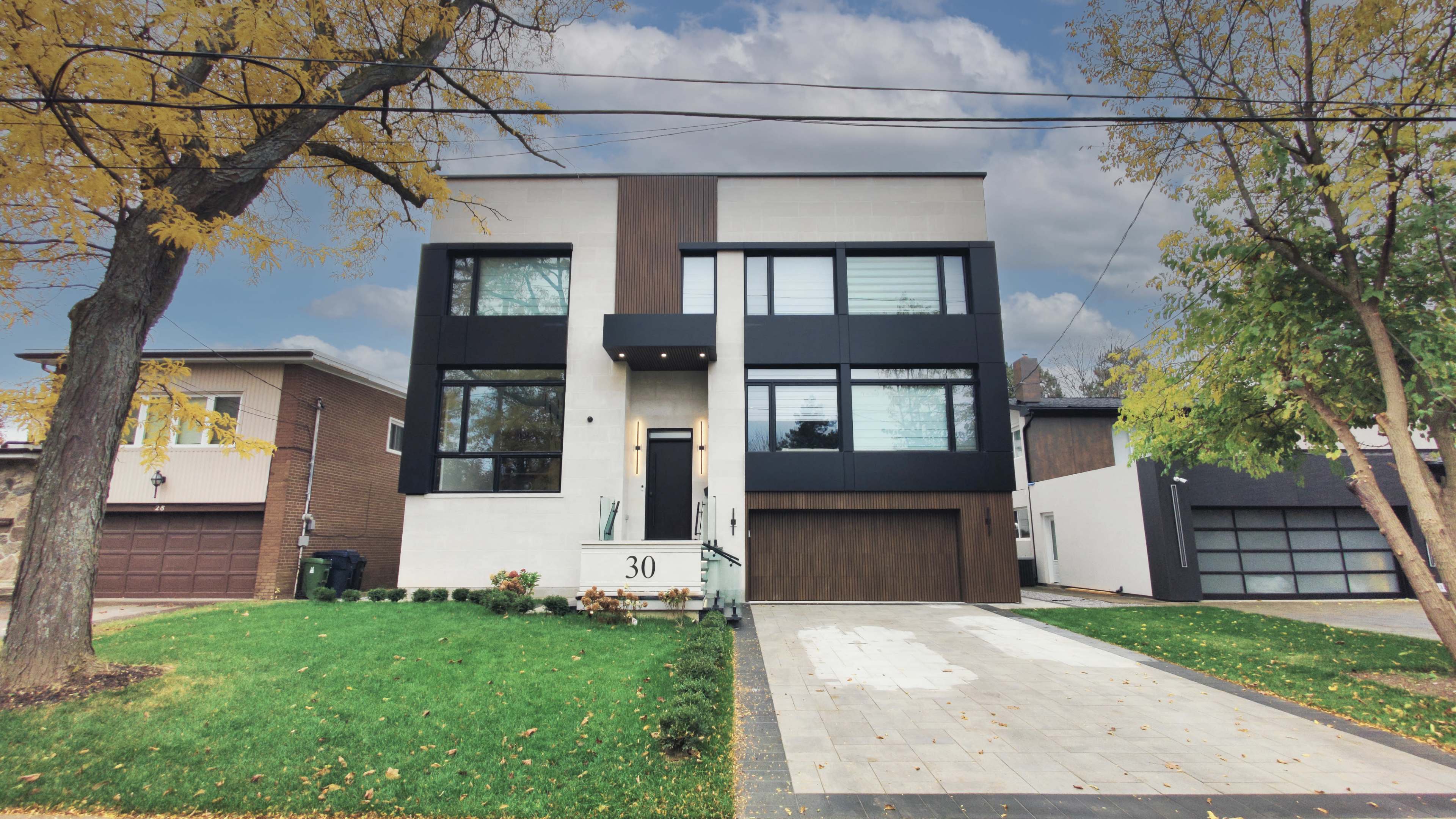 A modern two-story house with large windows and a mix of white and black panels is situated on a green lawn, with a concrete driveway leading to a garage on the side.