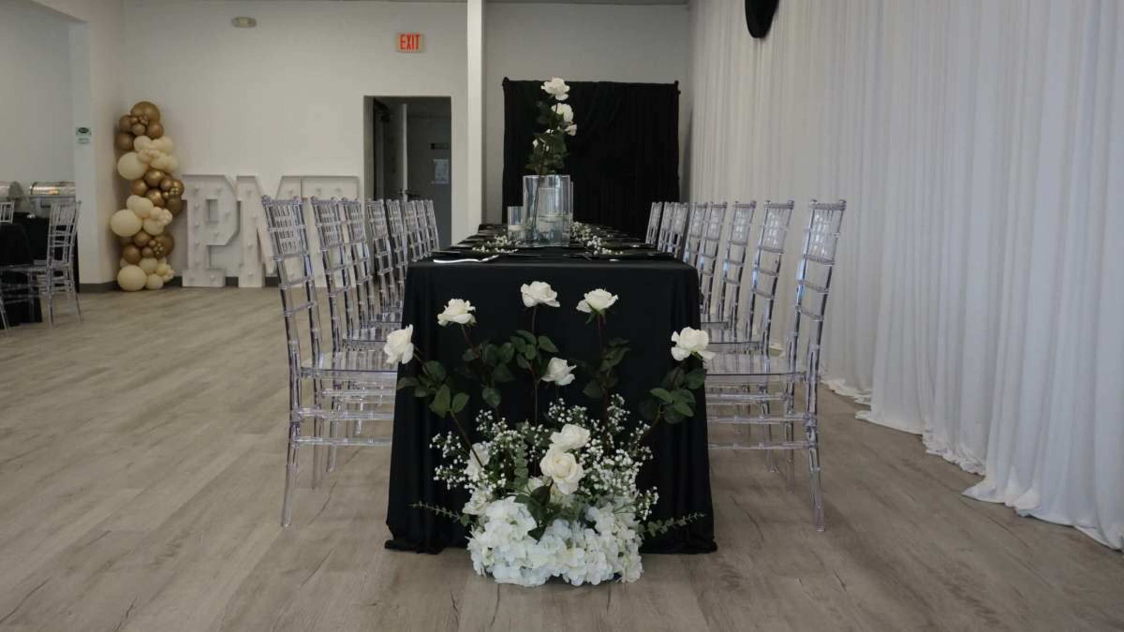 A long, elegantly set dining table with a black tablecloth, surrounded by transparent chairs, features floral arrangements of white roses and greenery at one end.
