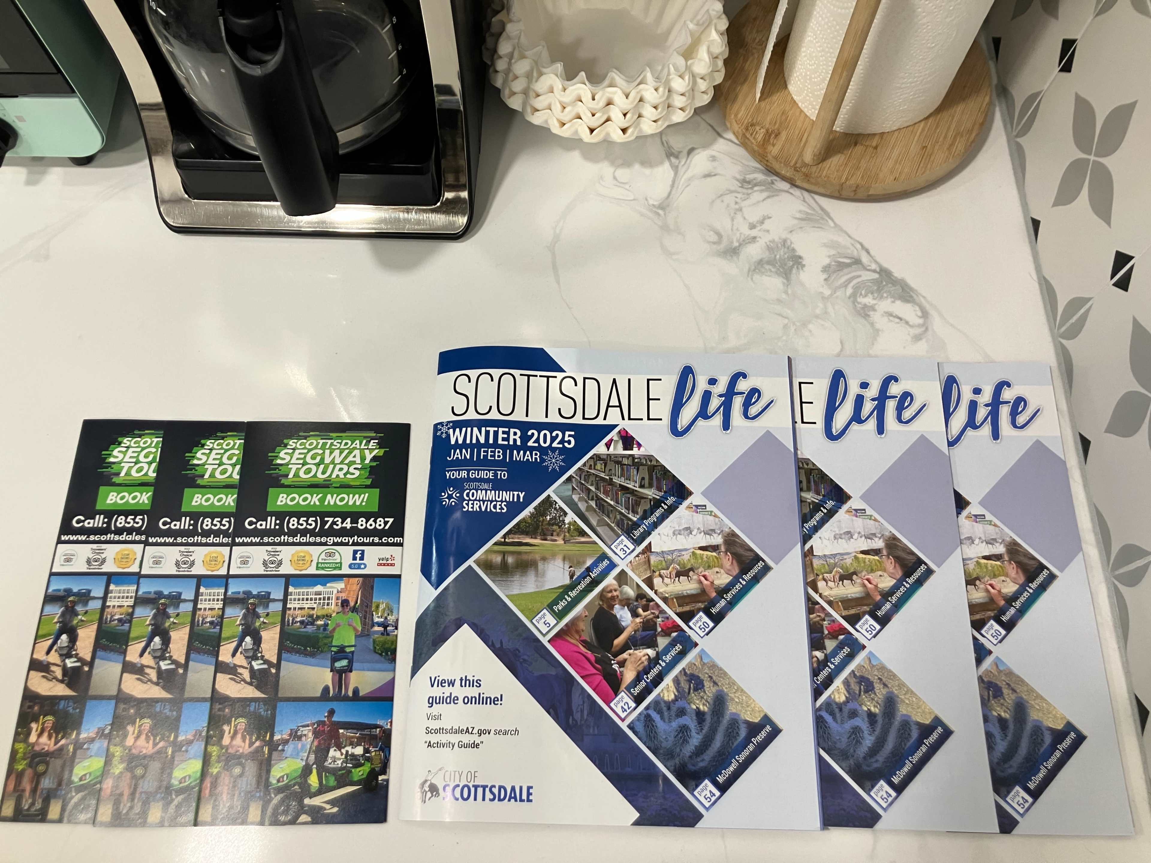 The image shows a countertop with brochures titled "SCOTTSDALE Life" and promotional materials for Segway tours, along with a coffee maker in the background.