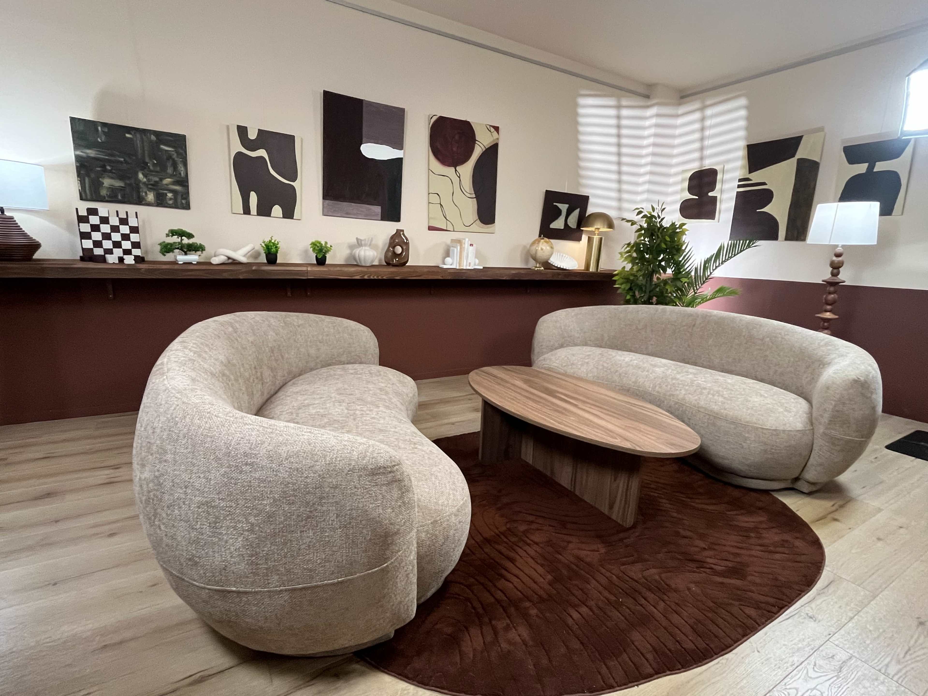 The image shows a cozy sitting area featuring two curved sofas around a round wooden coffee table, with abstract artwork on the walls and plants for decoration.