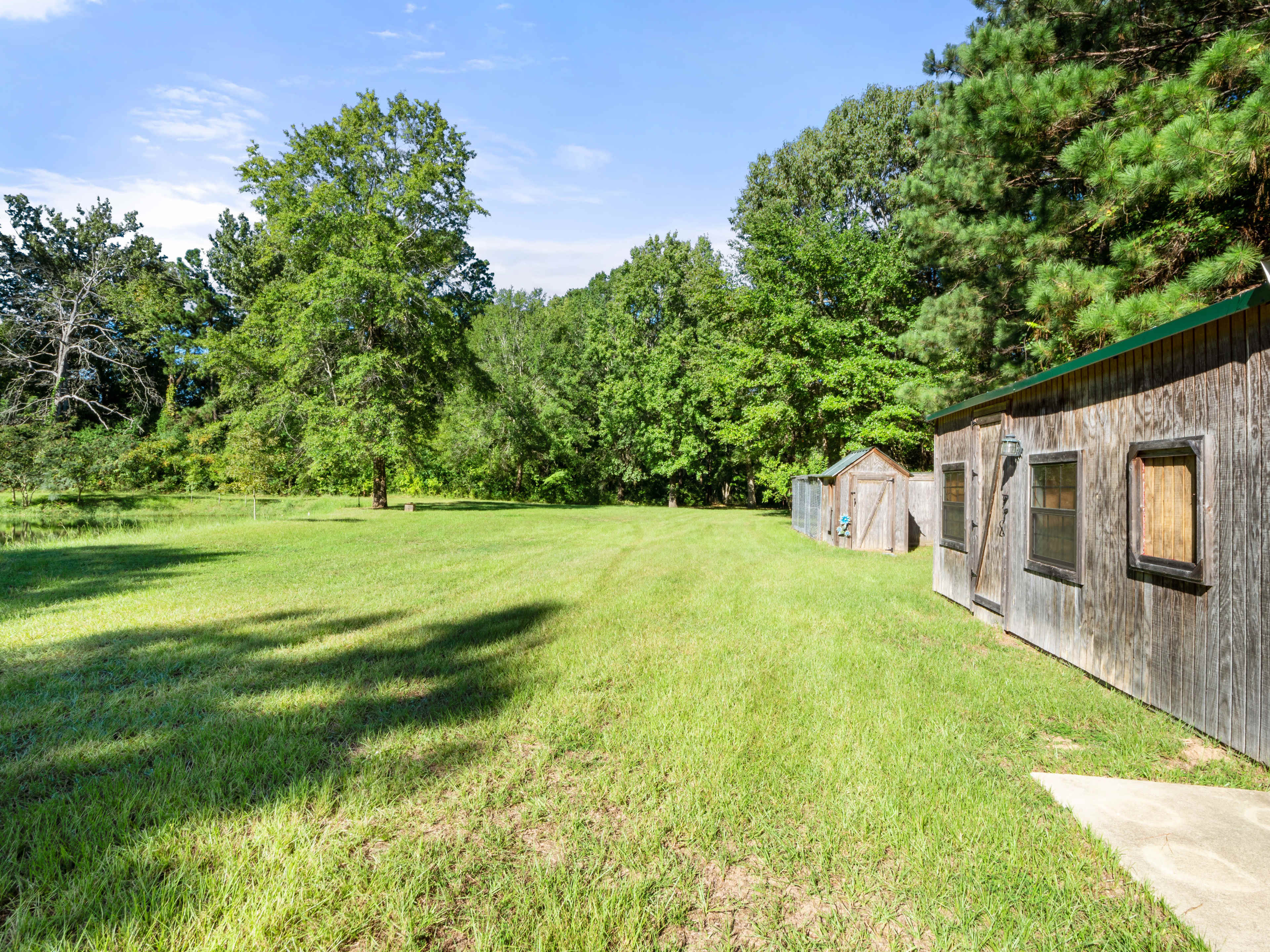 A wooden shed sits beside a grassy field surrounded by trees under a clear blue sky.