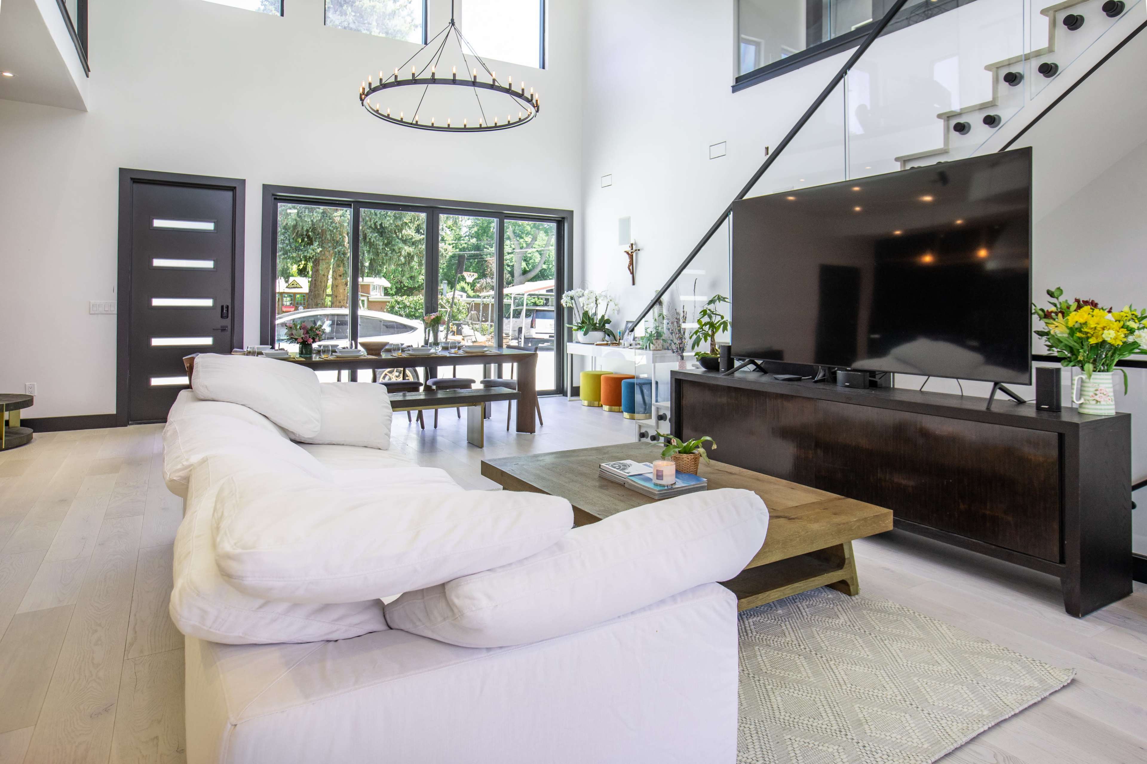 A modern living room featuring a white sectional sofa, a large flat-screen TV on a dark wooden console, and large windows that provide a view of the outdoors.