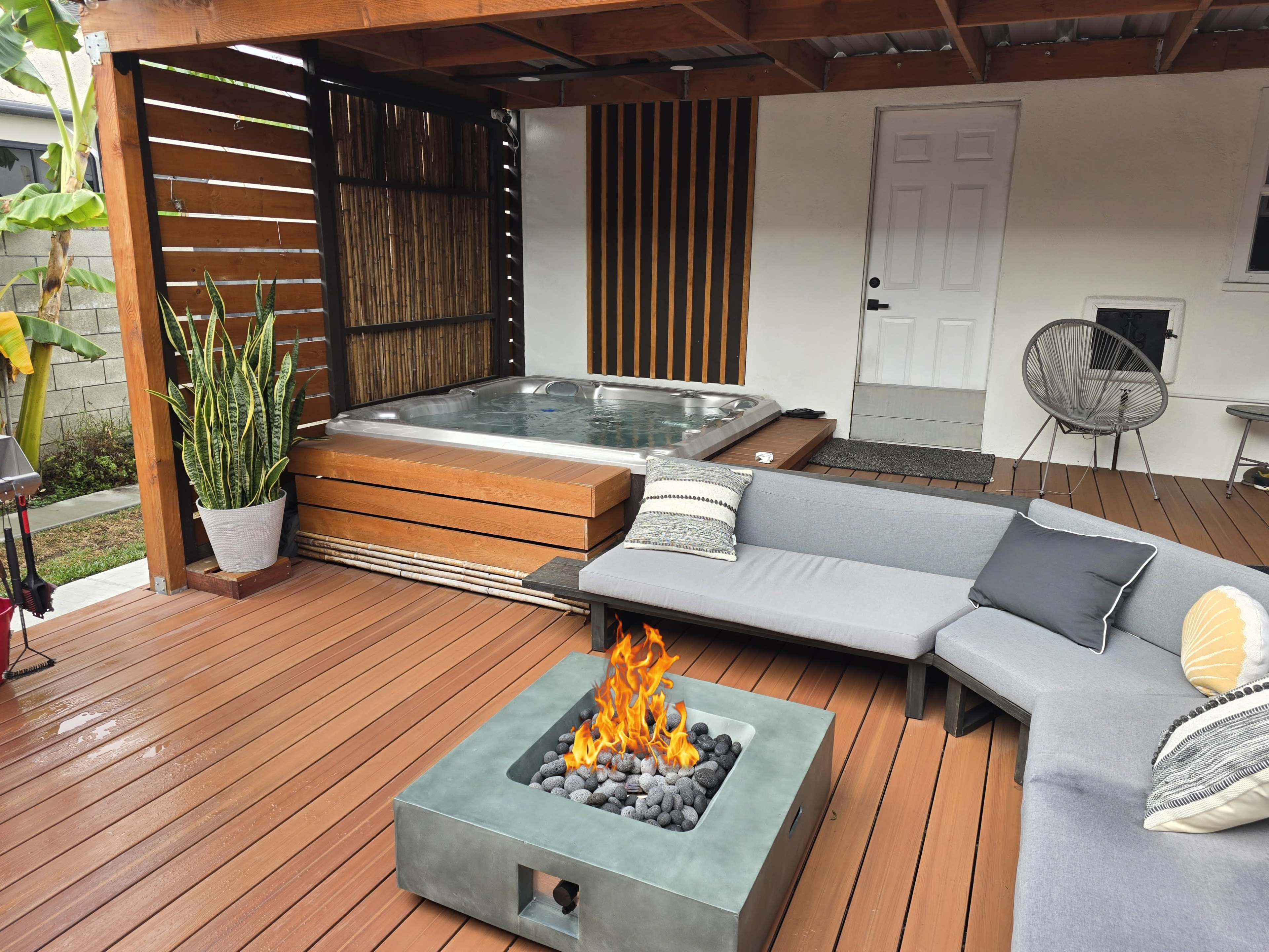 The image shows a modern outdoor patio with a hot tub, a seating area featuring a gray sectional sofa, and a square fire pit on wooden decking.