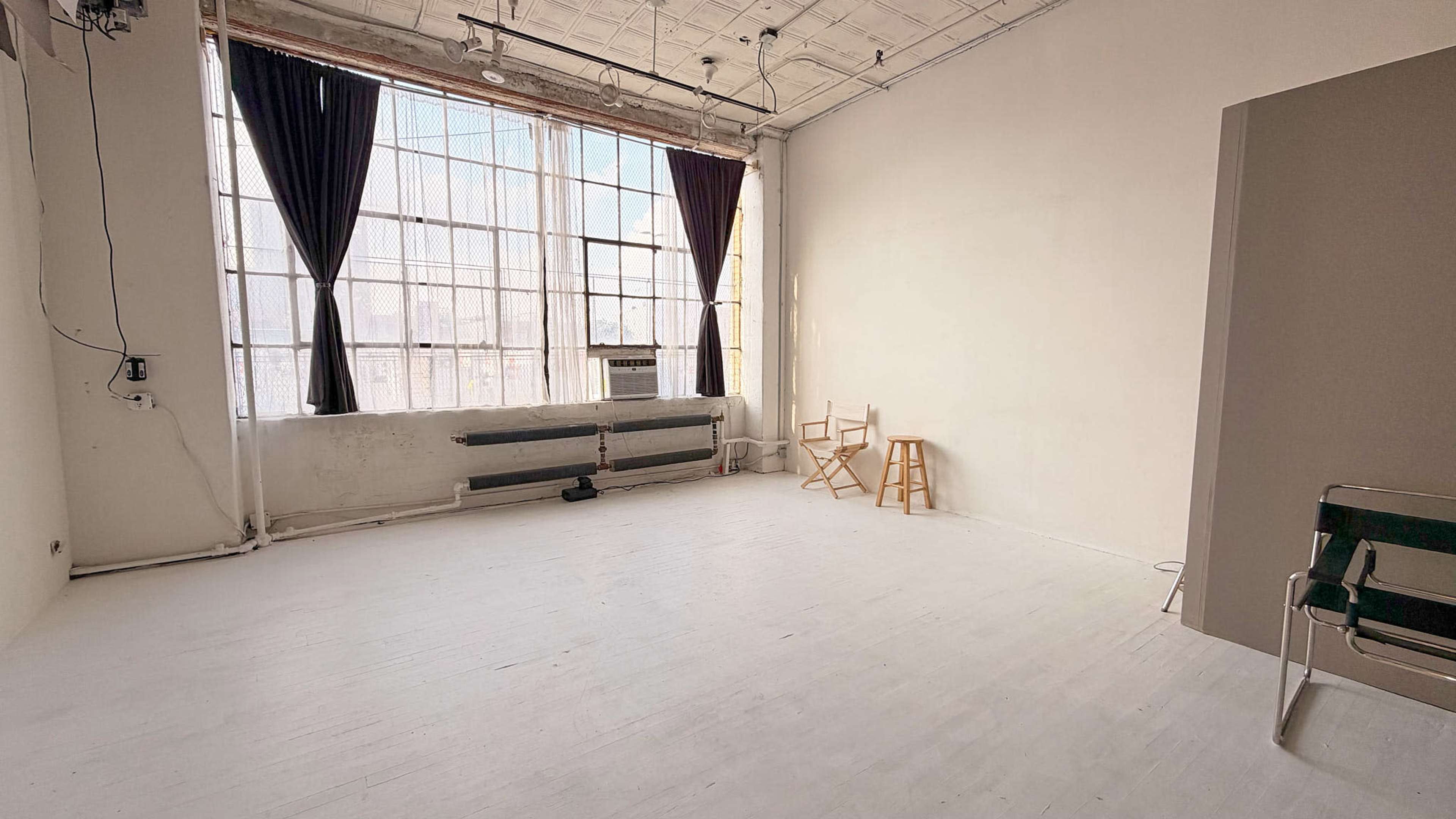 The image shows a spacious, empty studio with large windows, wooden flooring, and a few simple pieces of furniture.