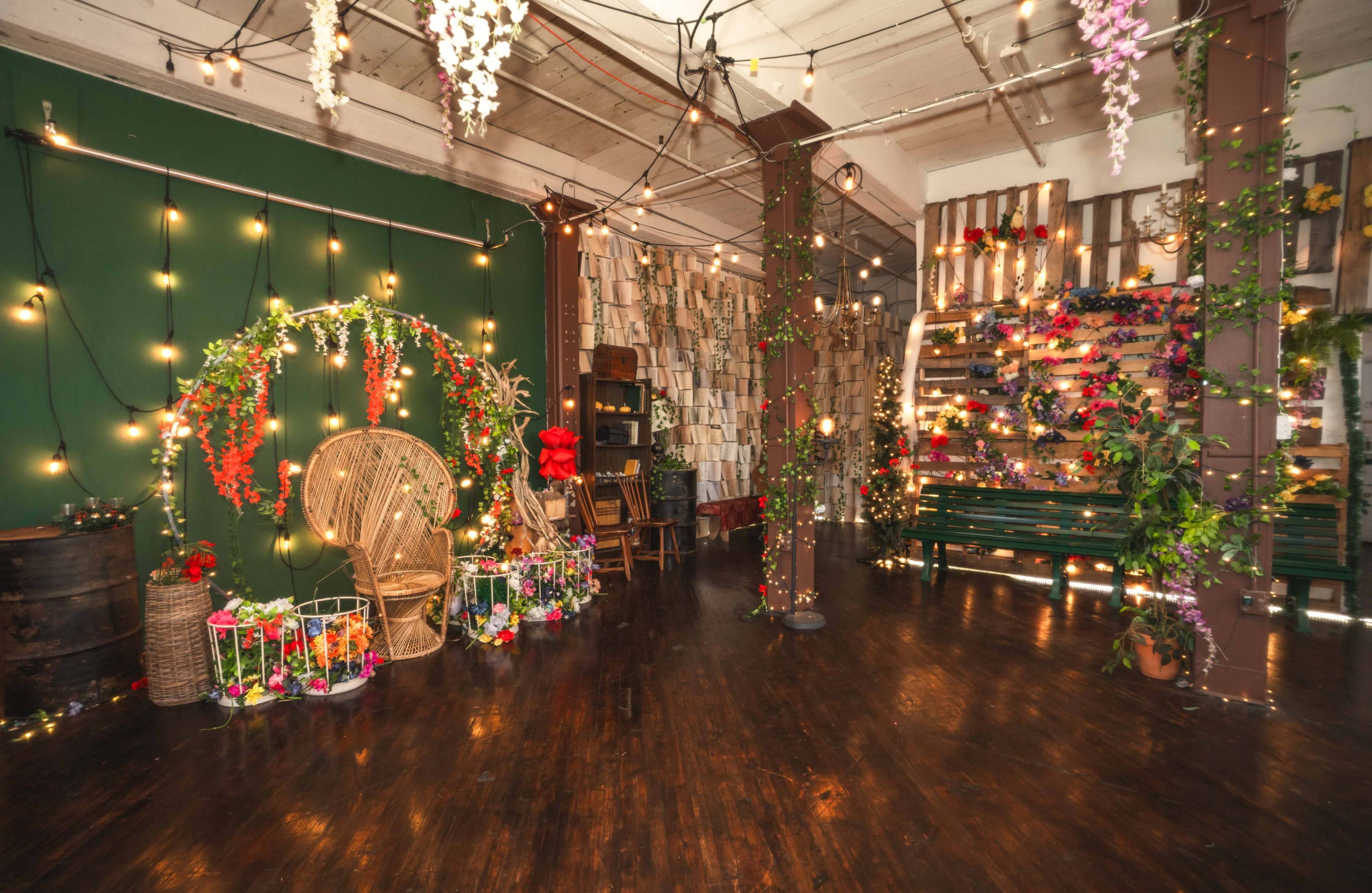 The image shows a decorated indoor space with floral arrangements, string lights, and various seating and display items, creating a vibrant atmosphere.