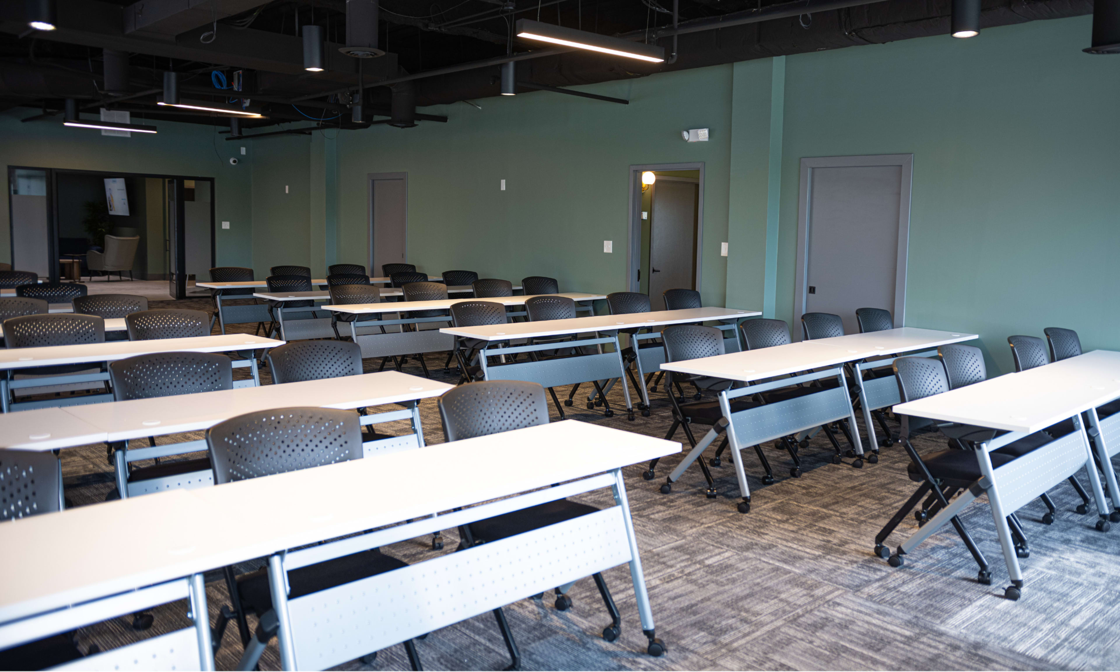 The image shows a classroom setting with multiple rows of tables and chairs arranged in a spacious, well-lit room.