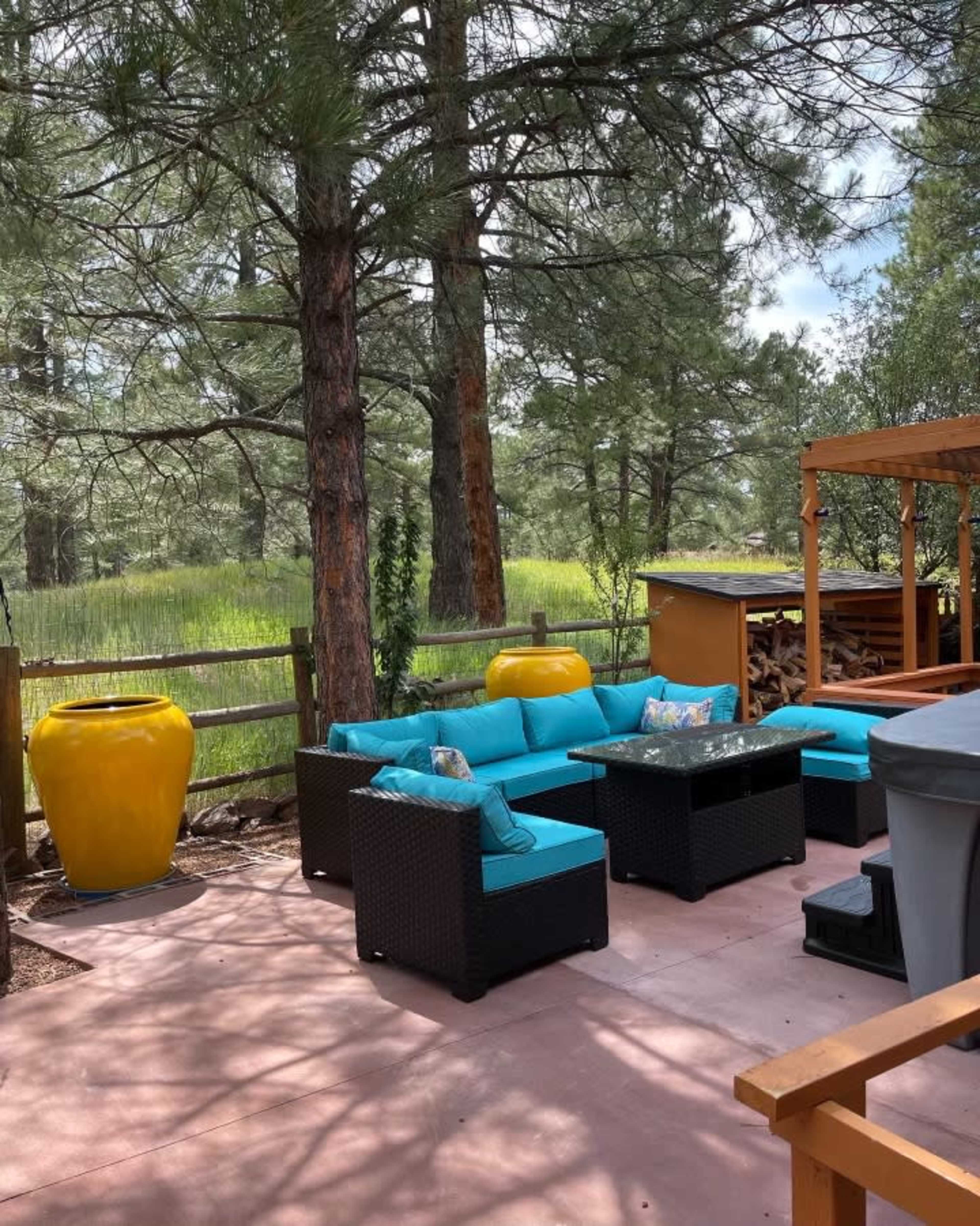 A patio area features turquoise furniture surrounded by tall pine trees and a wooden fence, with a yellow ceramic pot and a storage unit in the background.