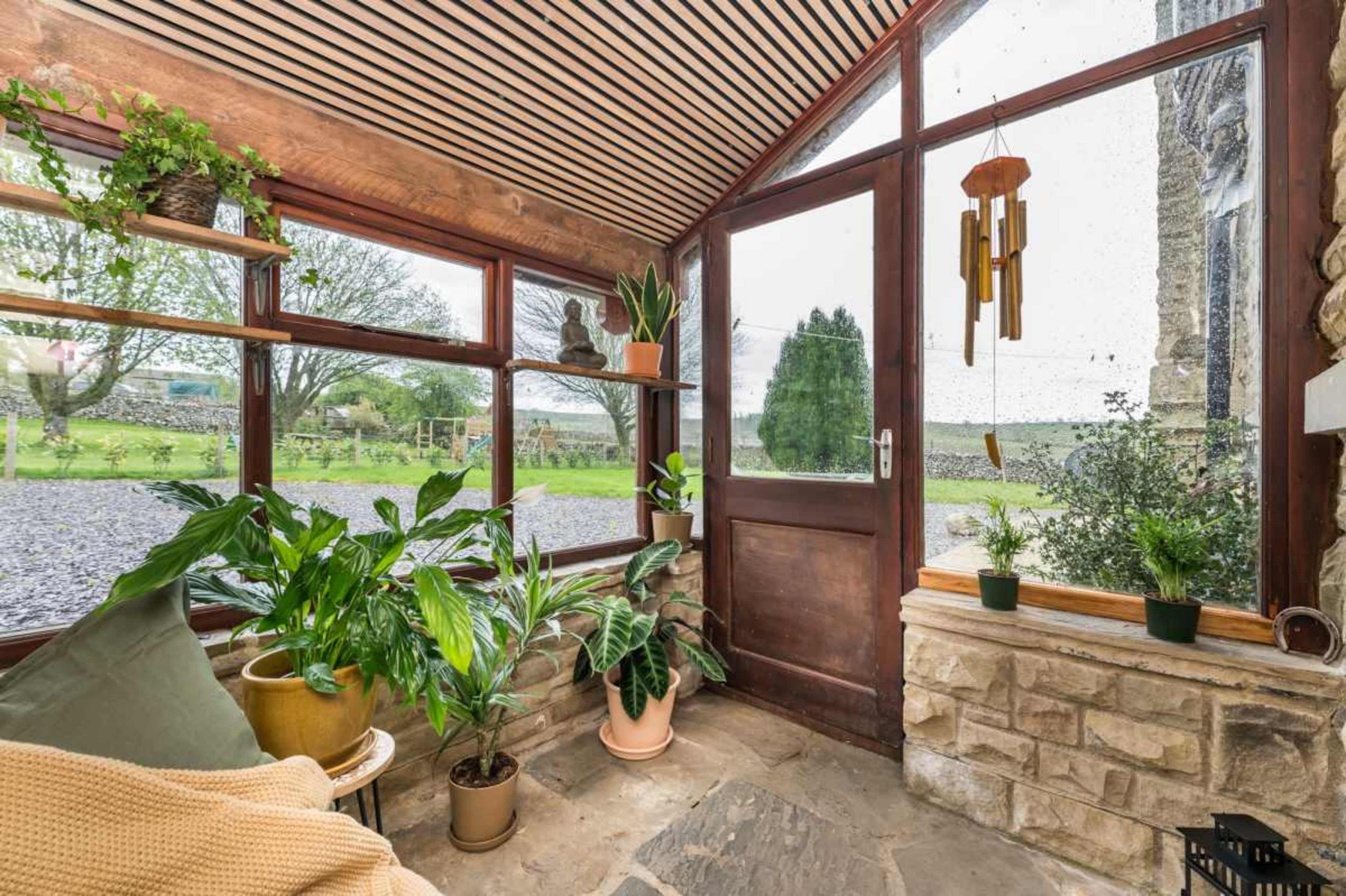A sunroom features a wooden structure with large windows, plants in pots, and a stone floor overlooking a grassy area outside.
