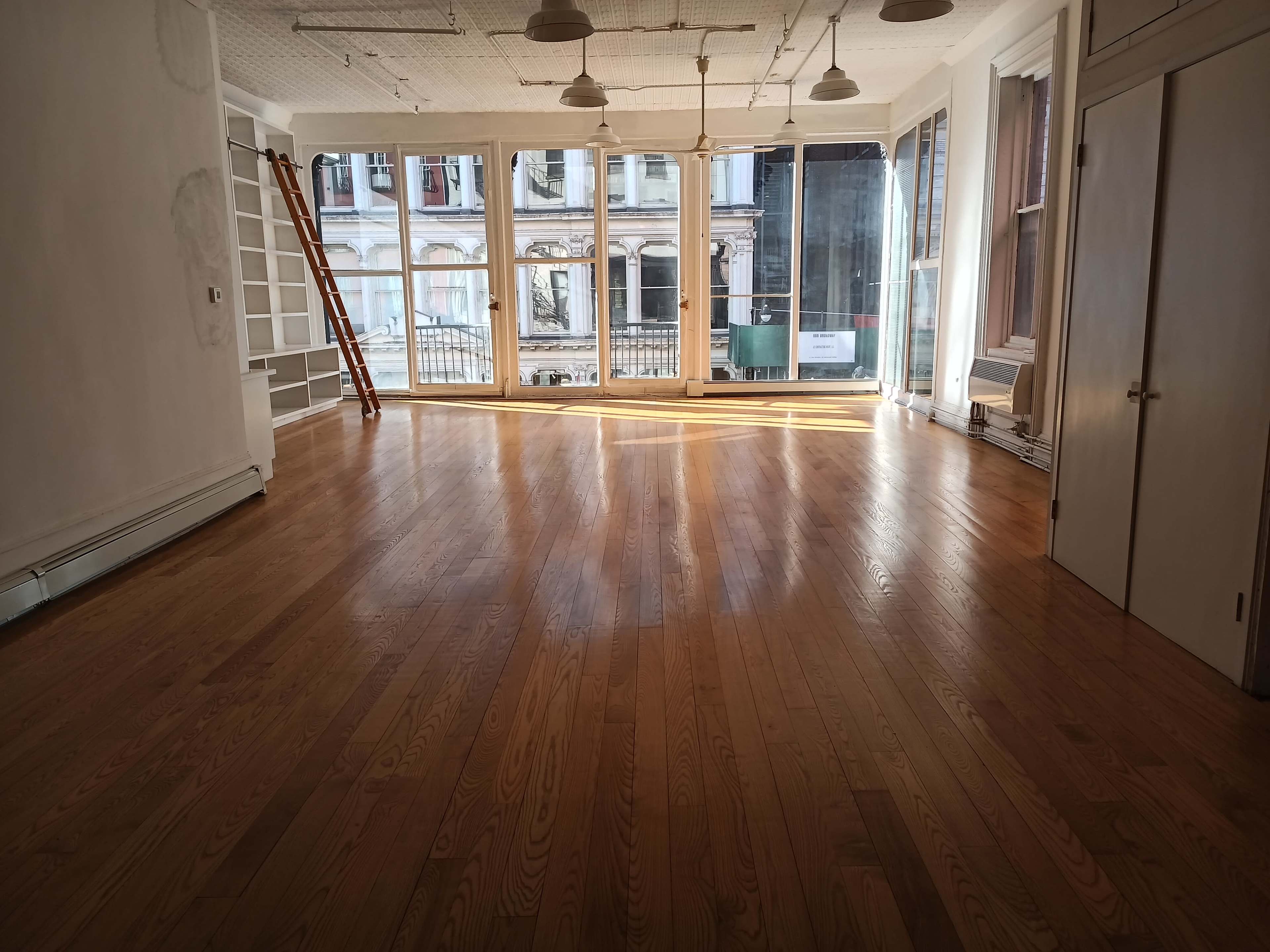 A spacious room with wooden floors and large windows letting in natural light, and there is a ladder leaning against a wall.