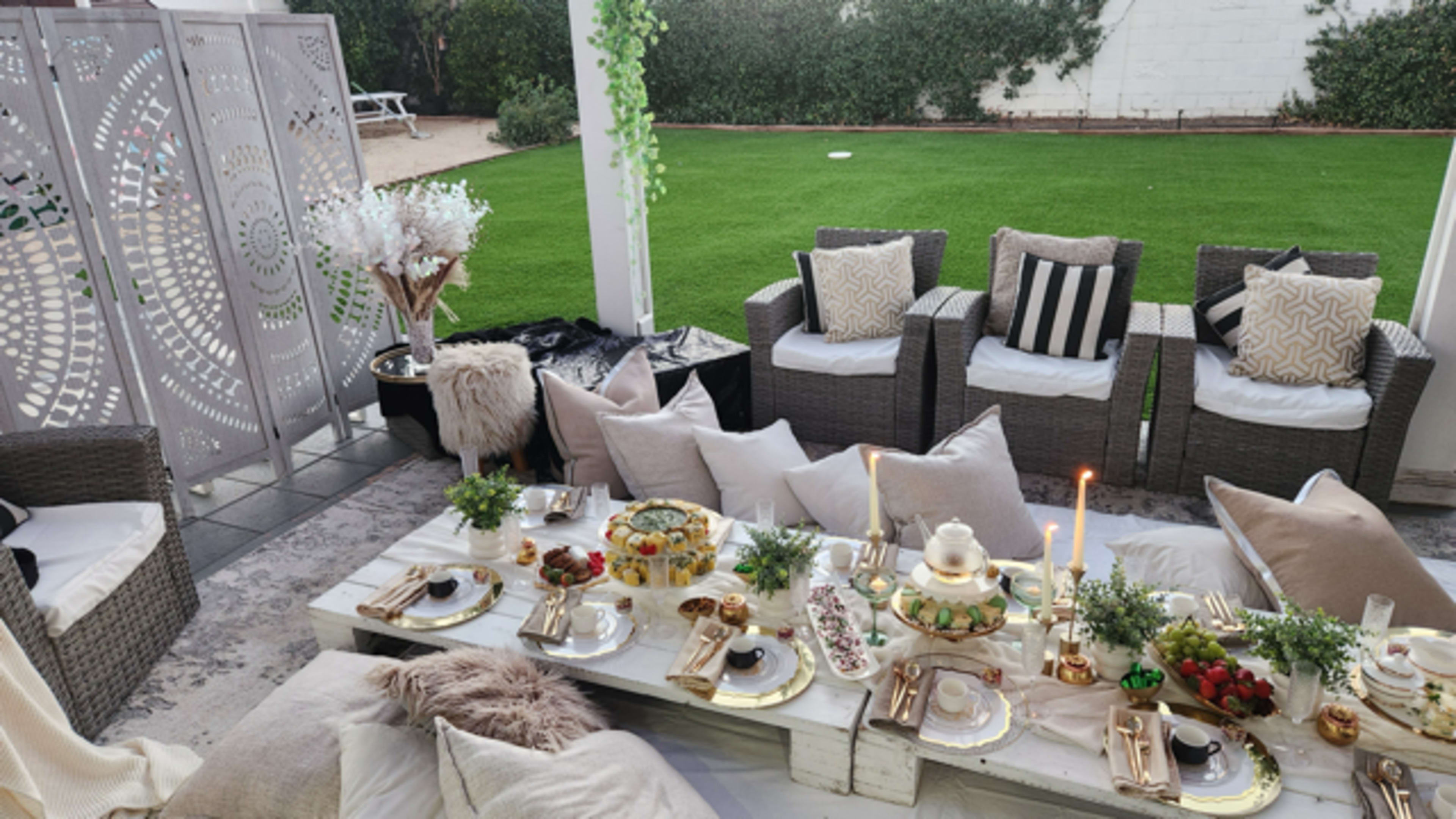 The image shows an outdoor seating area with a low table set for a meal, decorated with gold-detailed plates, candles, and greenery, surrounded by wicker chairs and plush cushions.