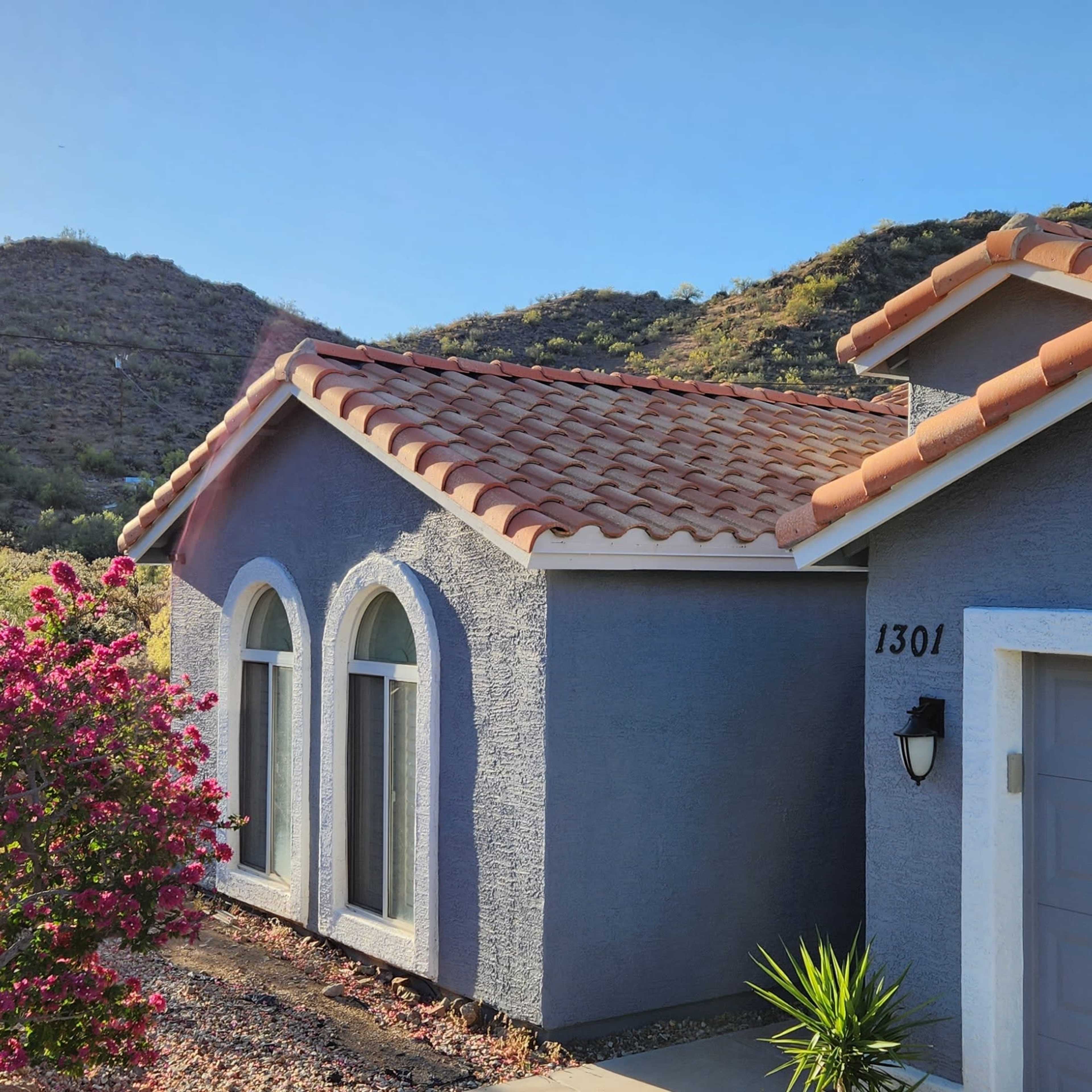 A blue house with arched windows and a tile roof is situated near a flowering bush and surrounded by hills.