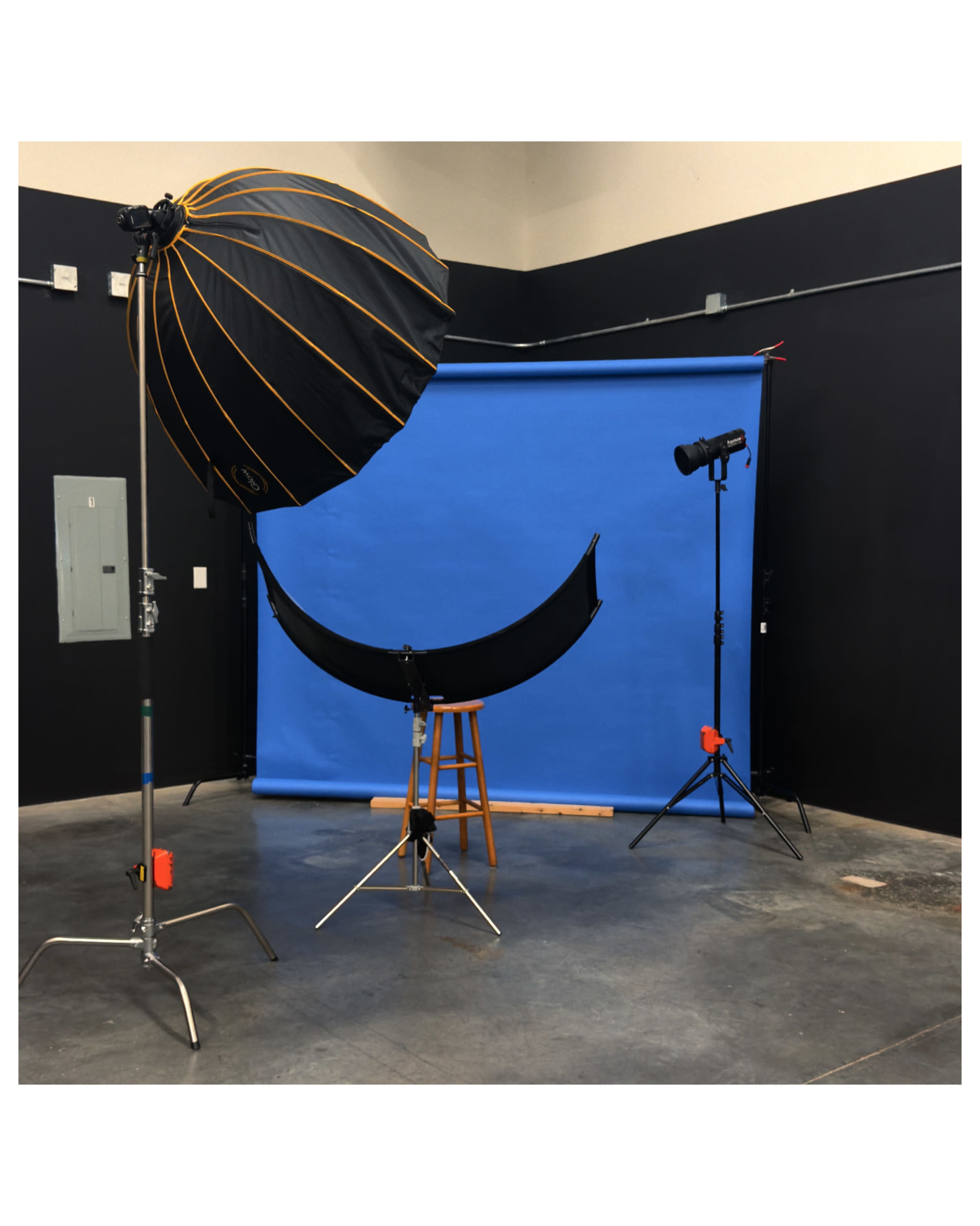 The image shows a photography studio setup featuring a blue backdrop, a black umbrella light, a light stand, and a wooden stool.