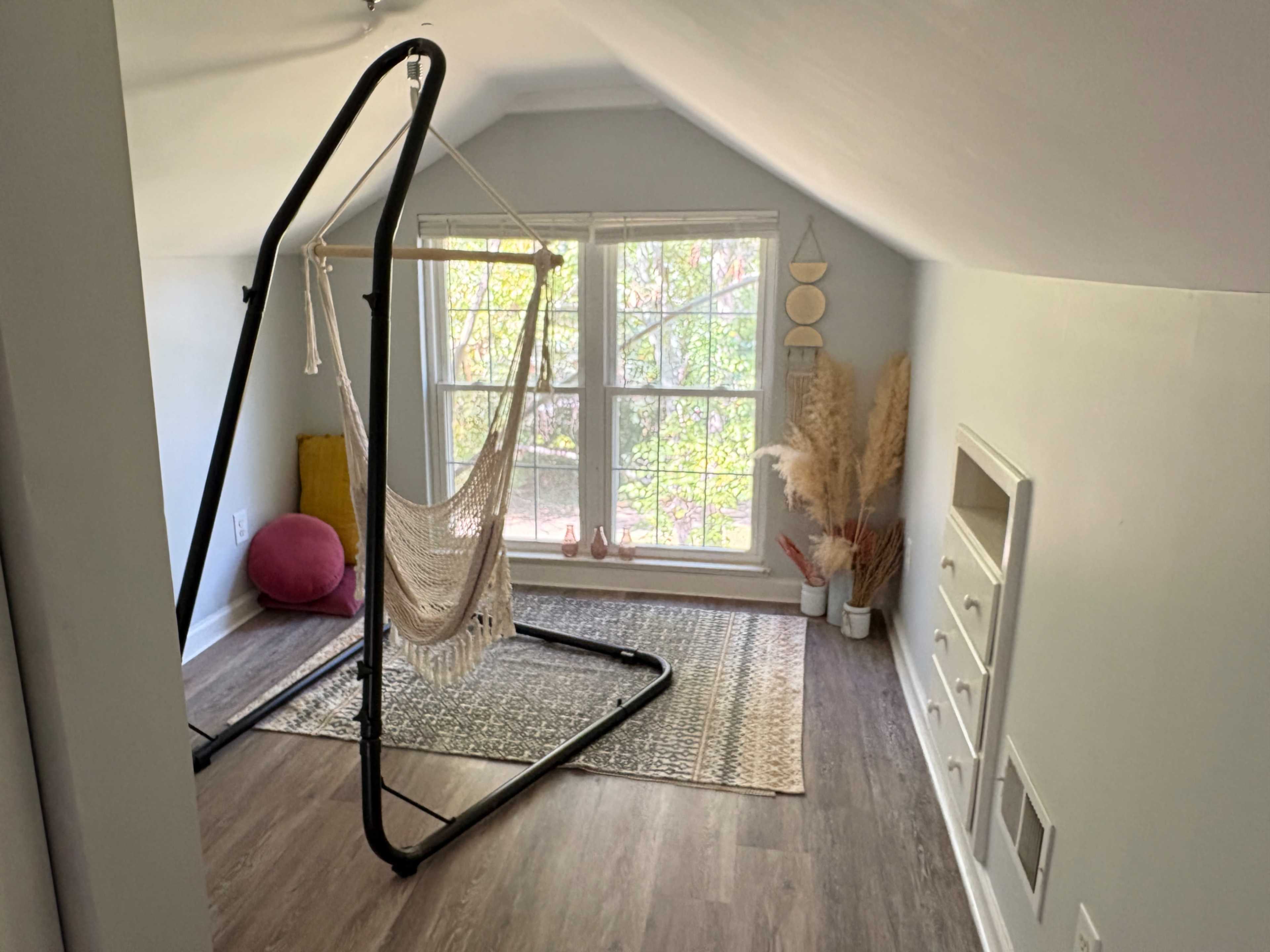 The room features a hanging hammock chair, a vibrant cushion, and large windows allowing natural light to illuminate the space.