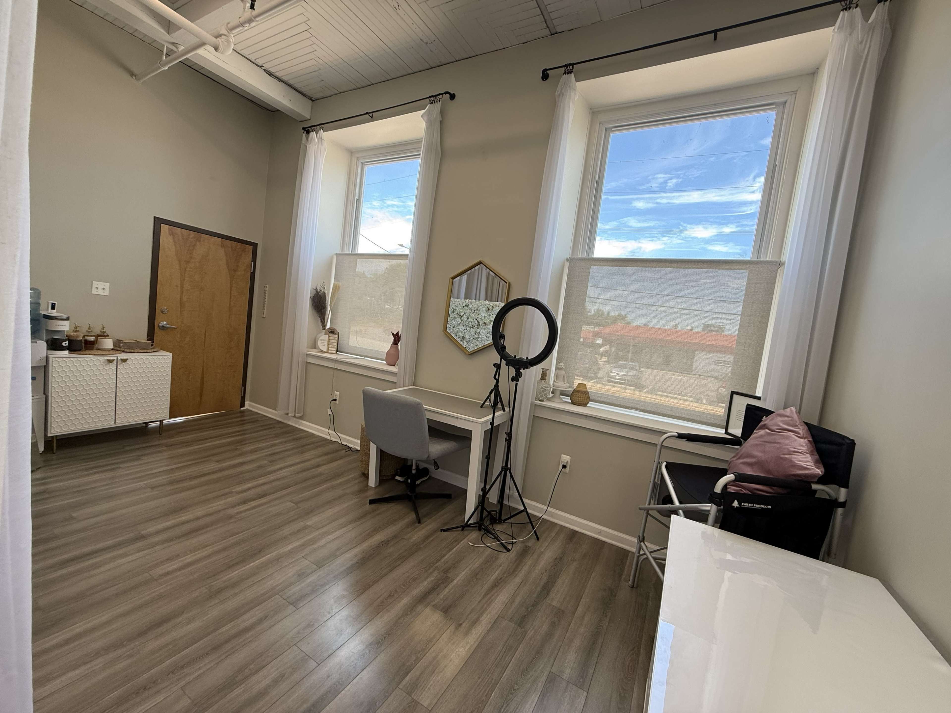 The room features two large windows, a desk with a chair, a ring light, and a cabinet against the wall.