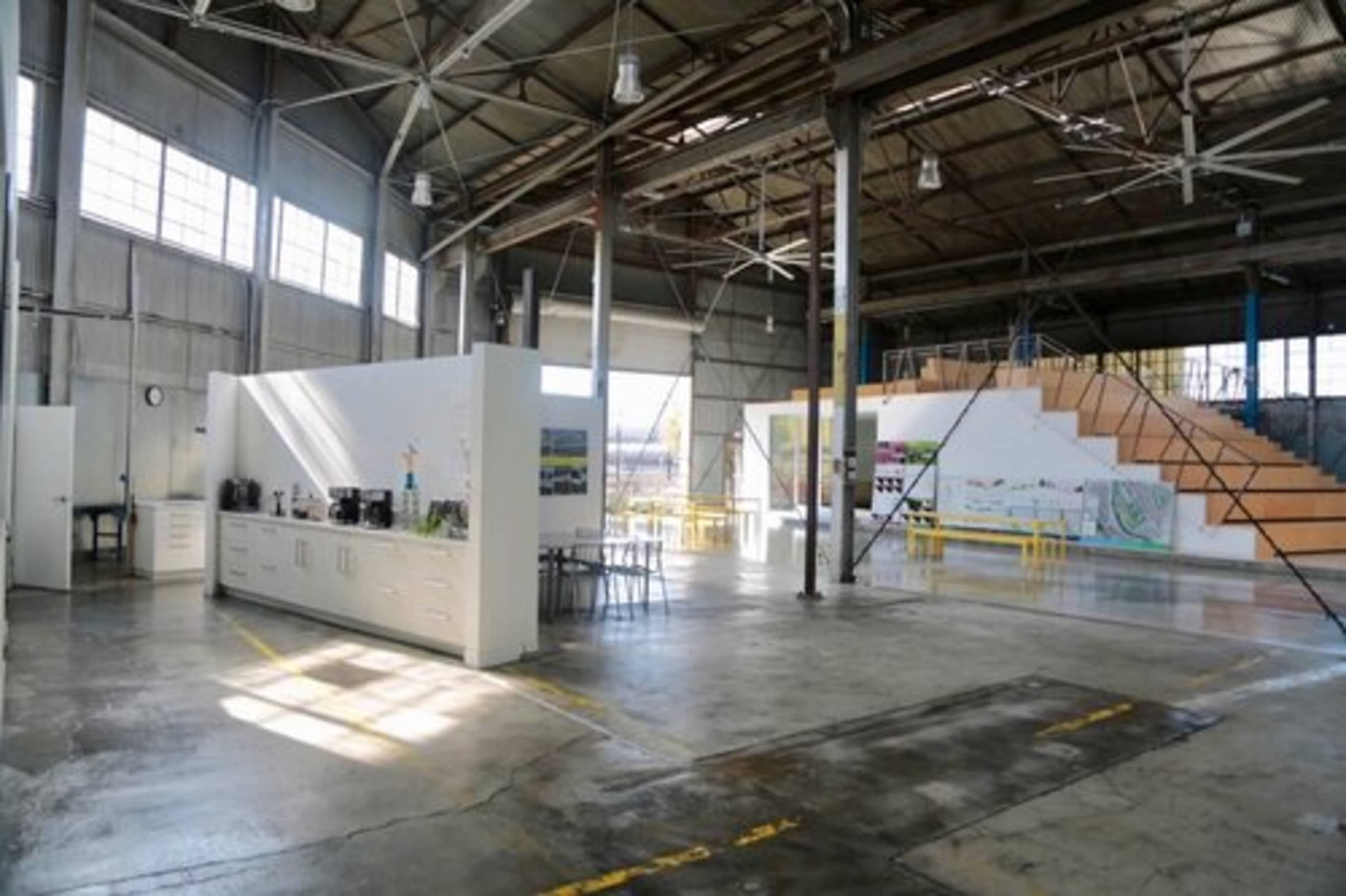 The image shows an industrial-style open-plan space with a kitchenette area, a set of stairs, and large windows allowing natural light to enter.