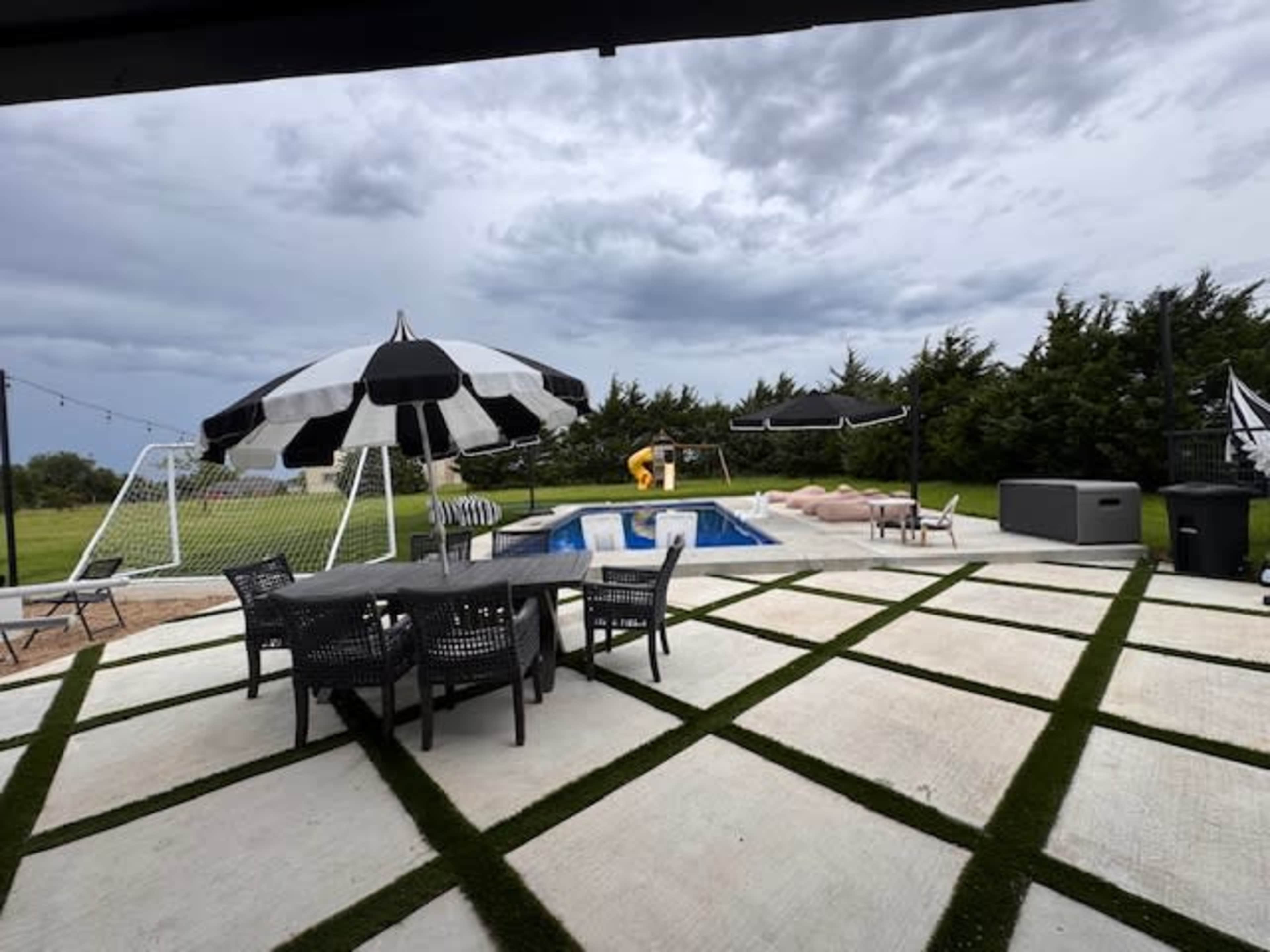 The image shows a patio area with a dining table, black and white umbrellas, a swimming pool, and a grassy lawn in the background.
