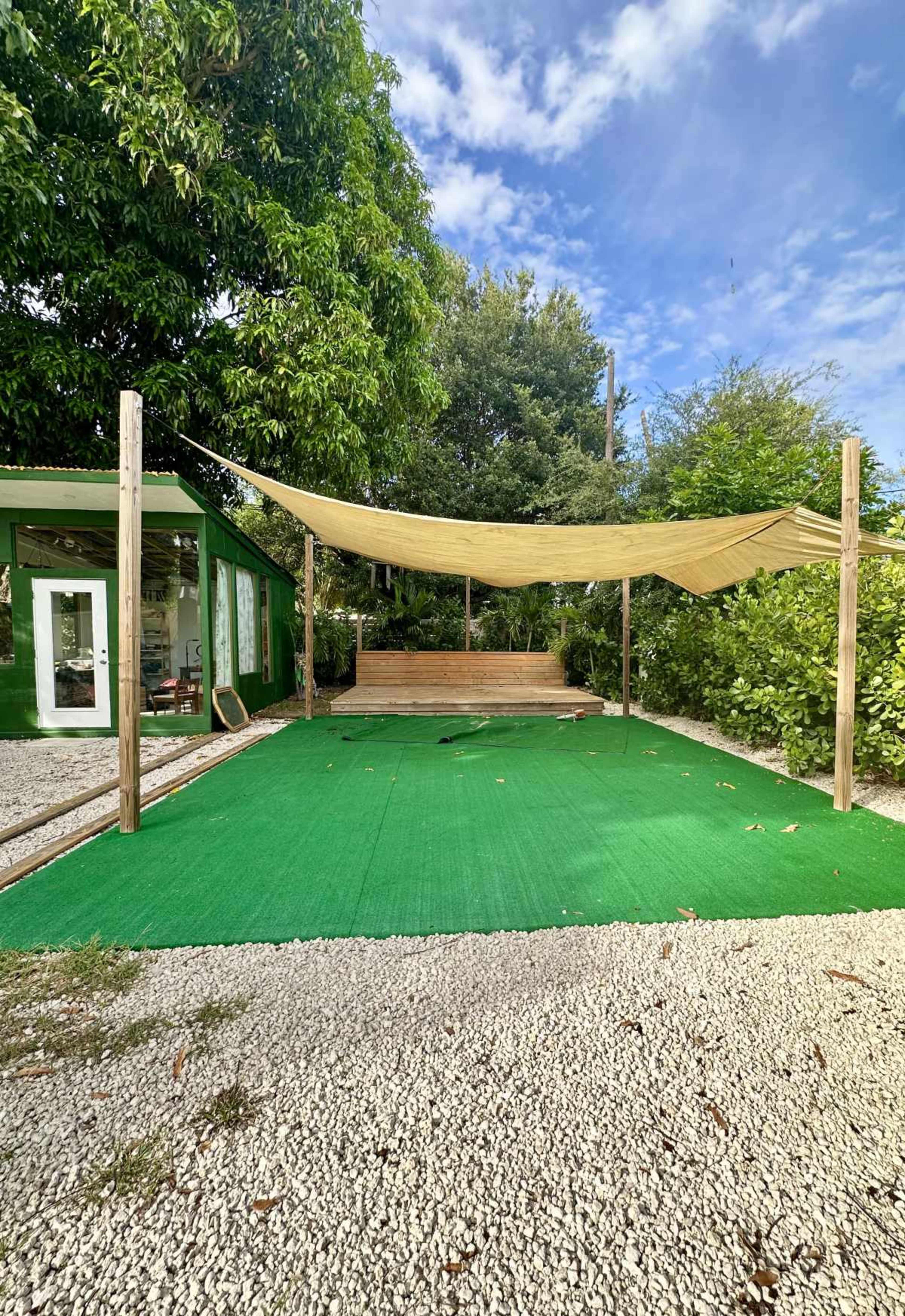 The image shows an outdoor area with a green artificial turf surface, a wooden seating space under a large shade sail, and a backdrop of greenery and a green structure.