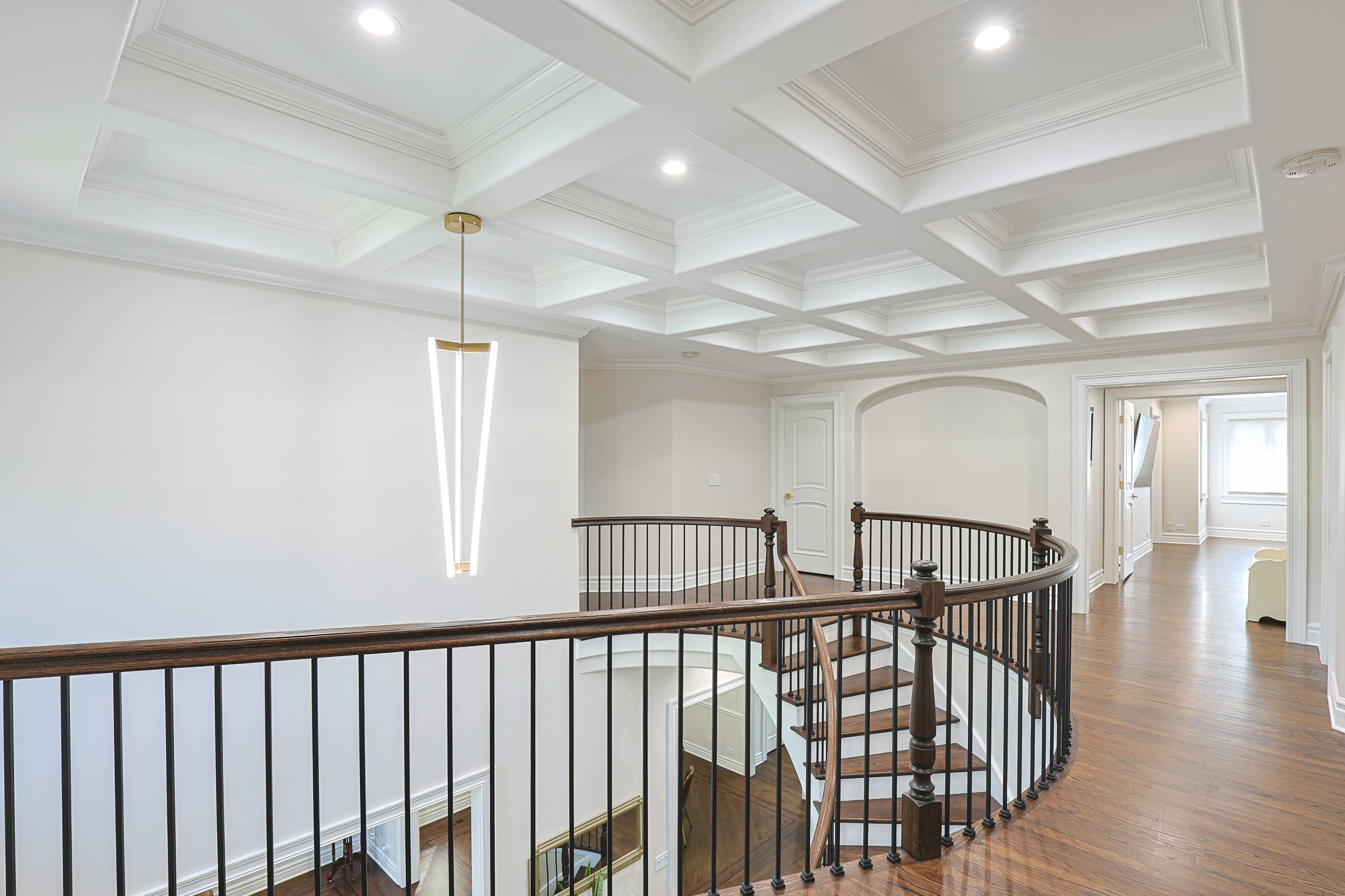 A well-lit, modern hallway featuring a coffered ceiling, a wooden railing staircase, and a contemporary light fixture.