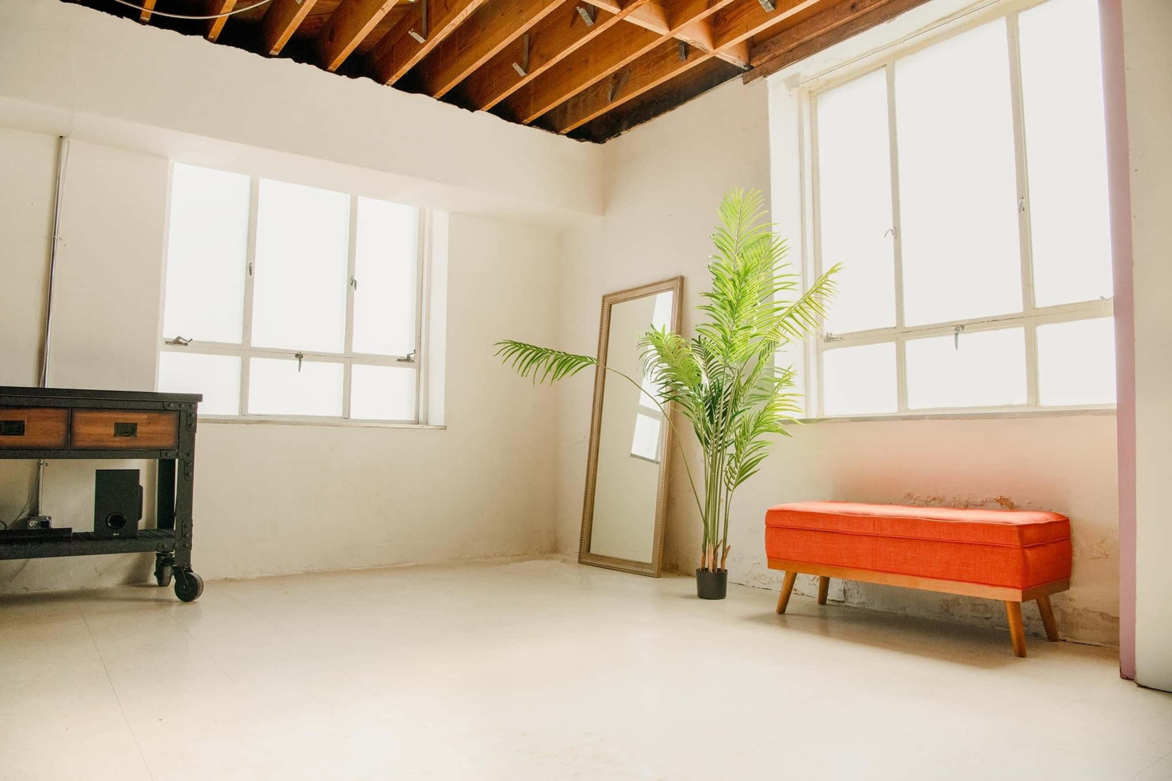 A minimally furnished room with large windows, a tall mirror, an orange upholstered bench, and a potted palm plant.