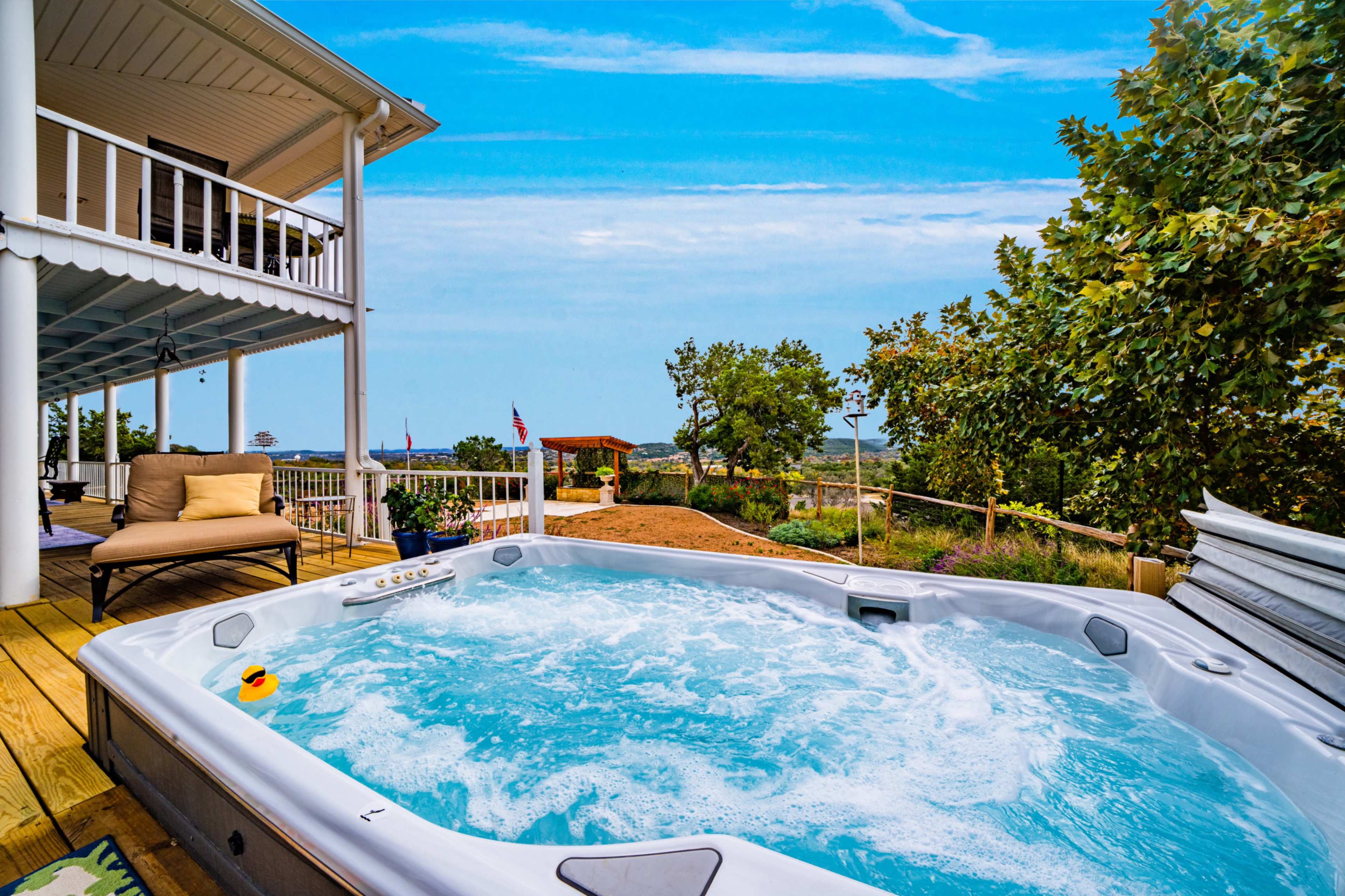 A hot tub bubbles on a deck overlooking a landscaped yard and distant trees under a blue sky.
