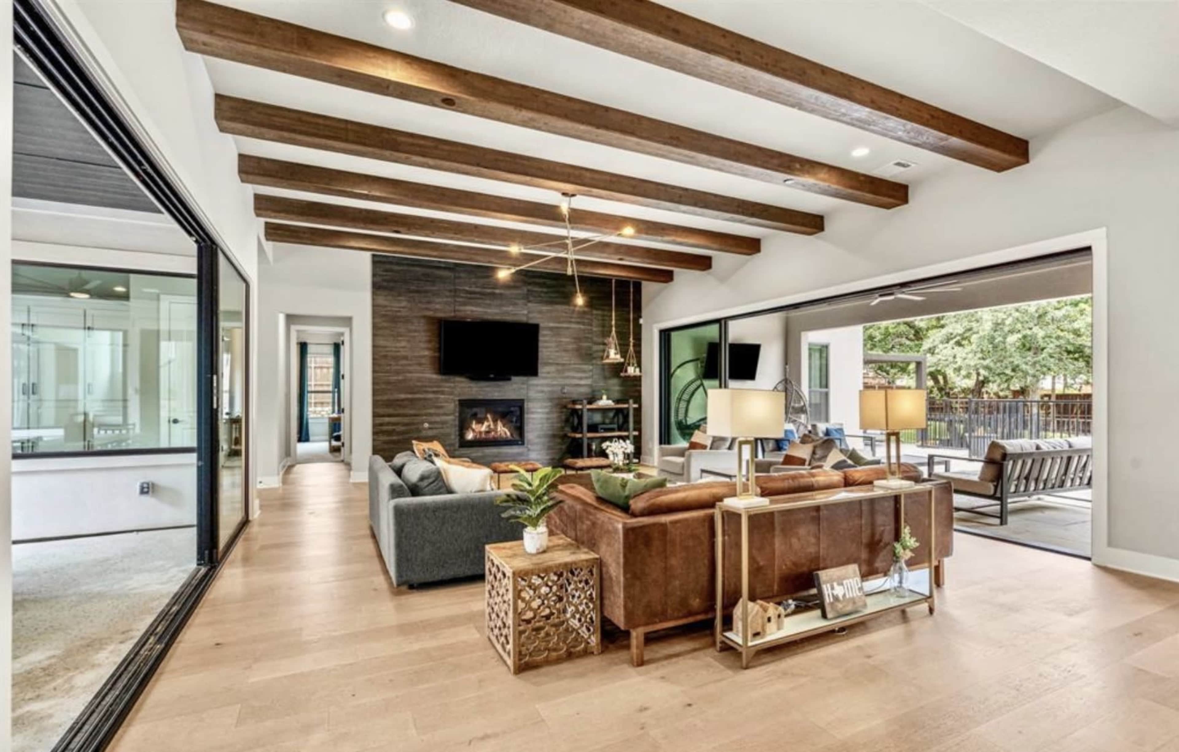 The image shows a modern living room with wooden beams, a fireplace, and a view of an outdoor patio through large sliding glass doors.