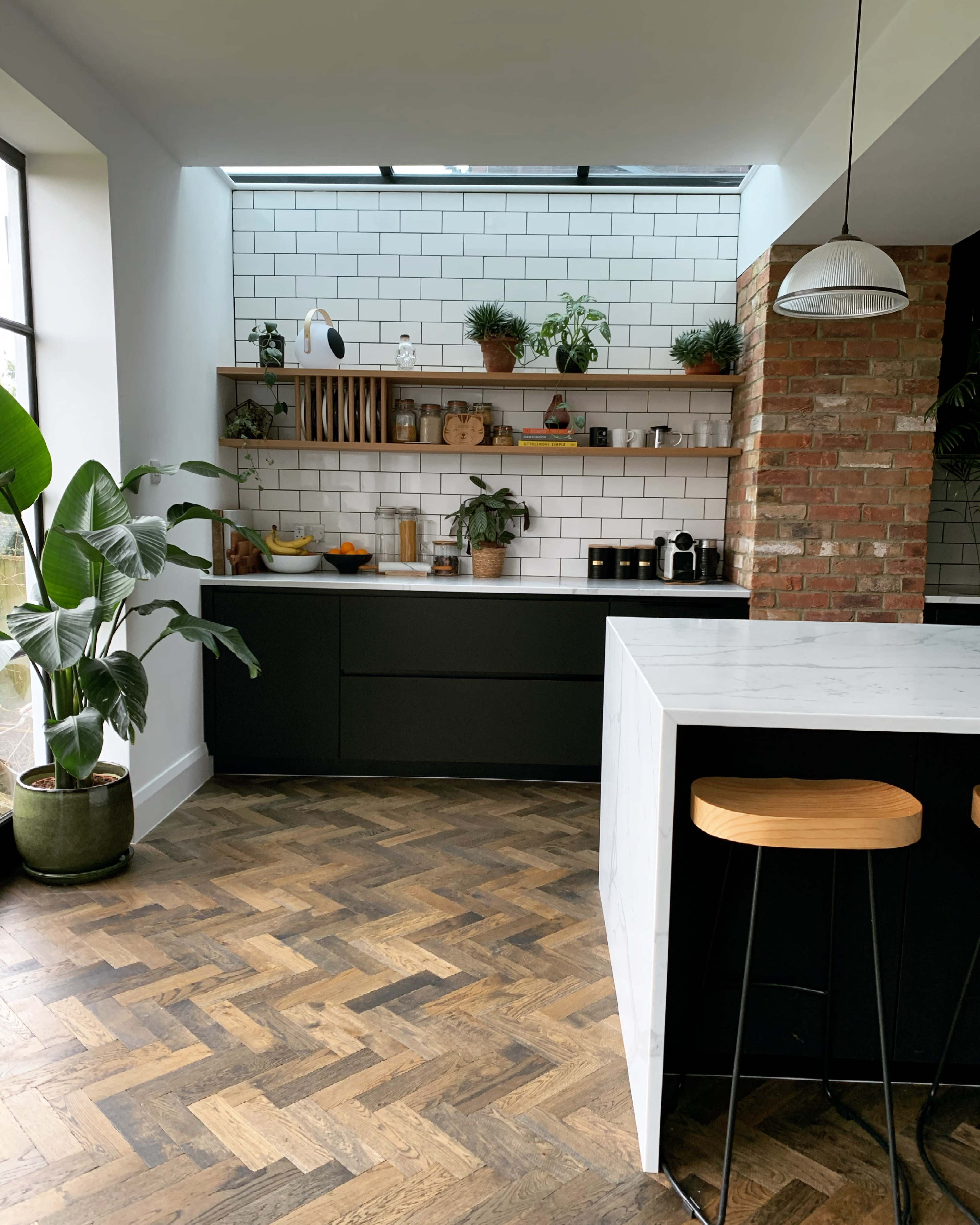 A modern kitchen features dark cabinets, a marble-top island with bar stools, and open shelving adorned with plants and ceramics against a backdrop of white subway tiles.