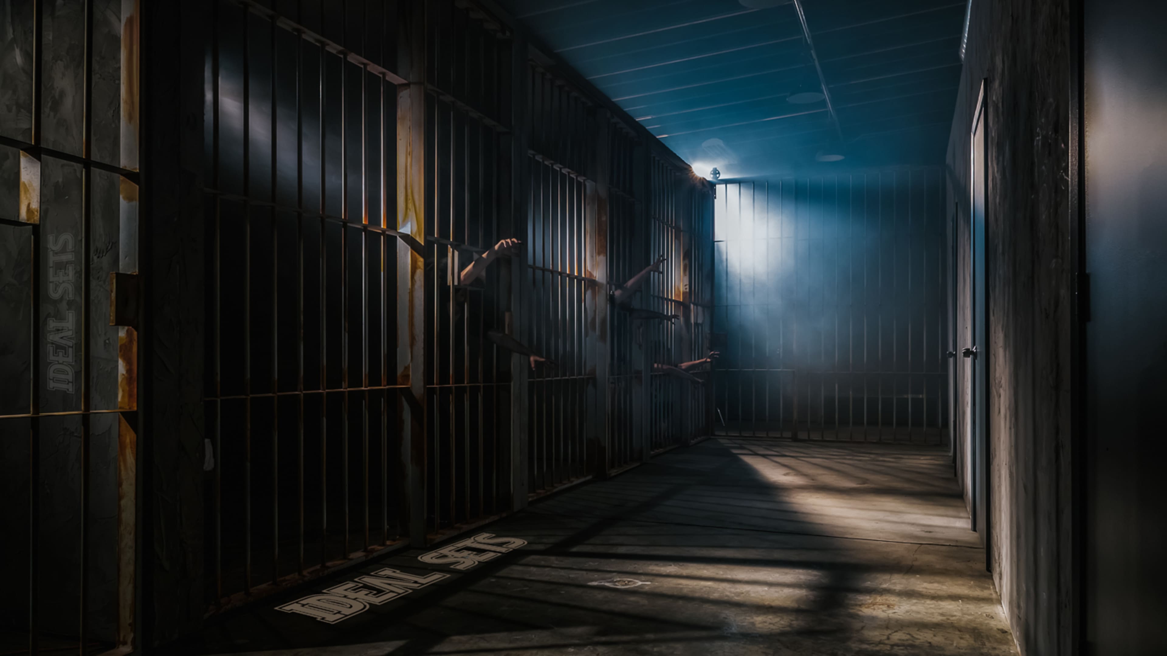 The image shows a dimly lit corridor with jail cells on one side, featuring shadowy hands reaching through the bars.
