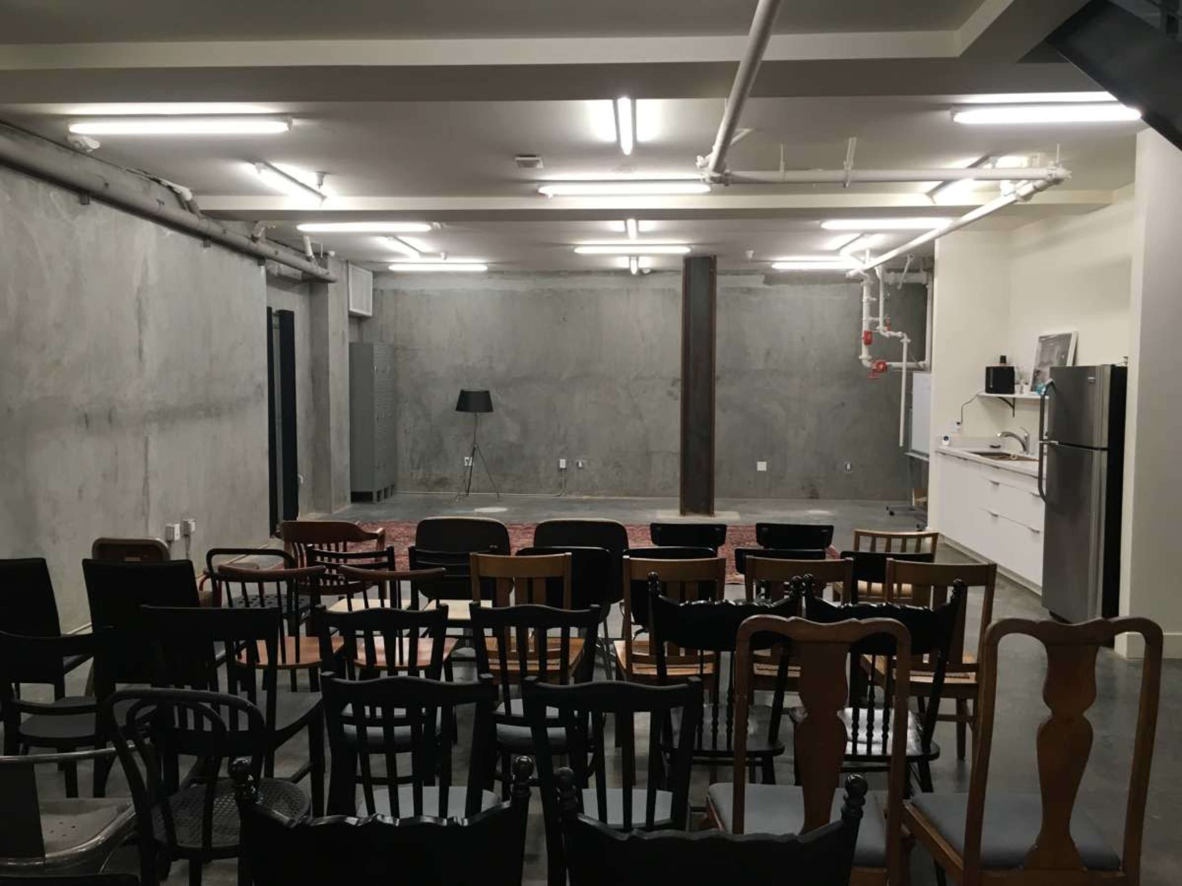 The image shows an empty room with a concrete wall, a row of wooden chairs facing a small stage area, and a kitchenette in the background.