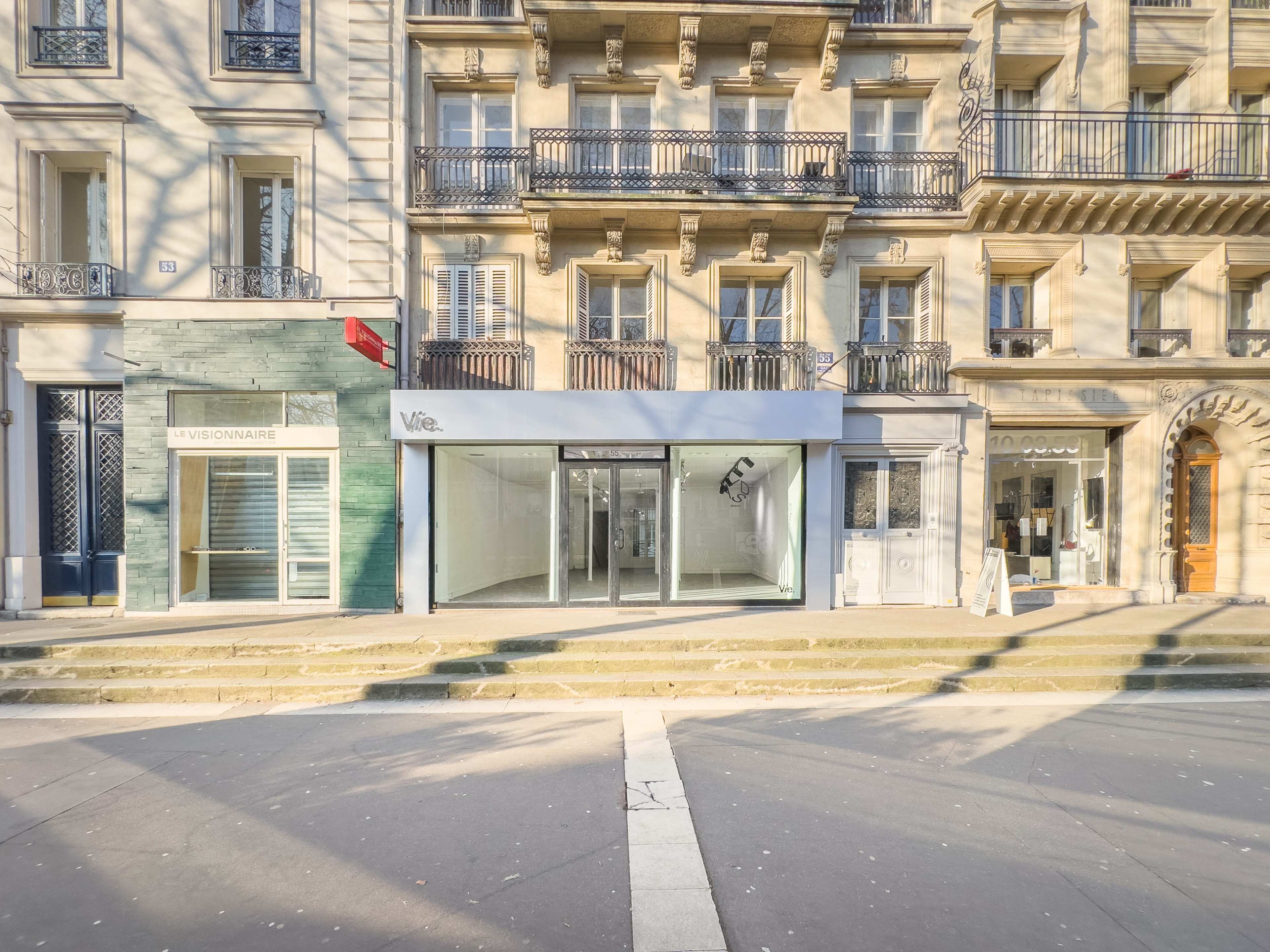 The image shows a street scene featuring several storefronts with distinct architectural styles, including a modern glass-front shop labeled "Vie" and a textured green facade nearby.