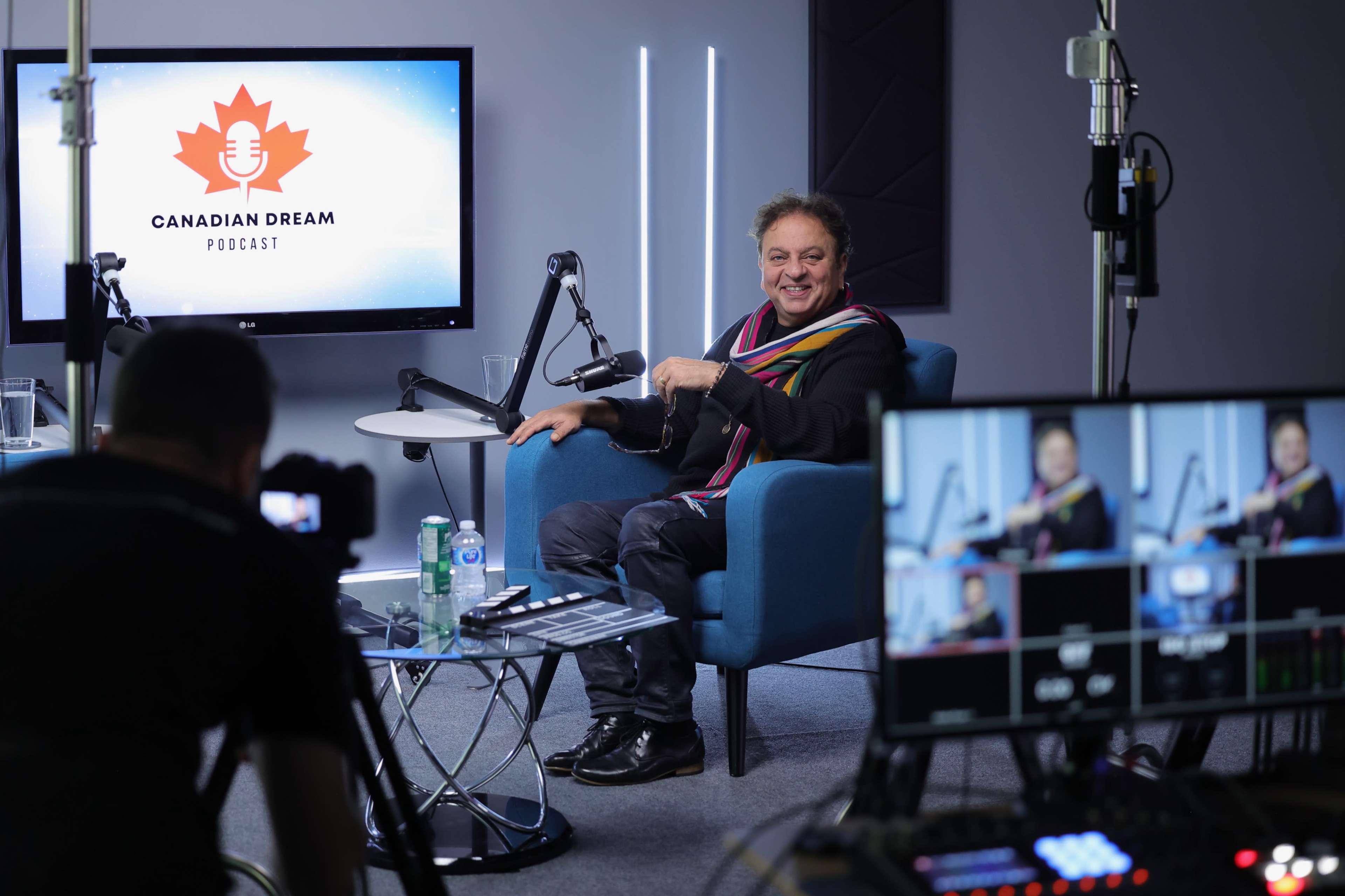 A man sits in a blue chair speaking during a recording of the "Canadian Dream Podcast" in a modern studio setup.