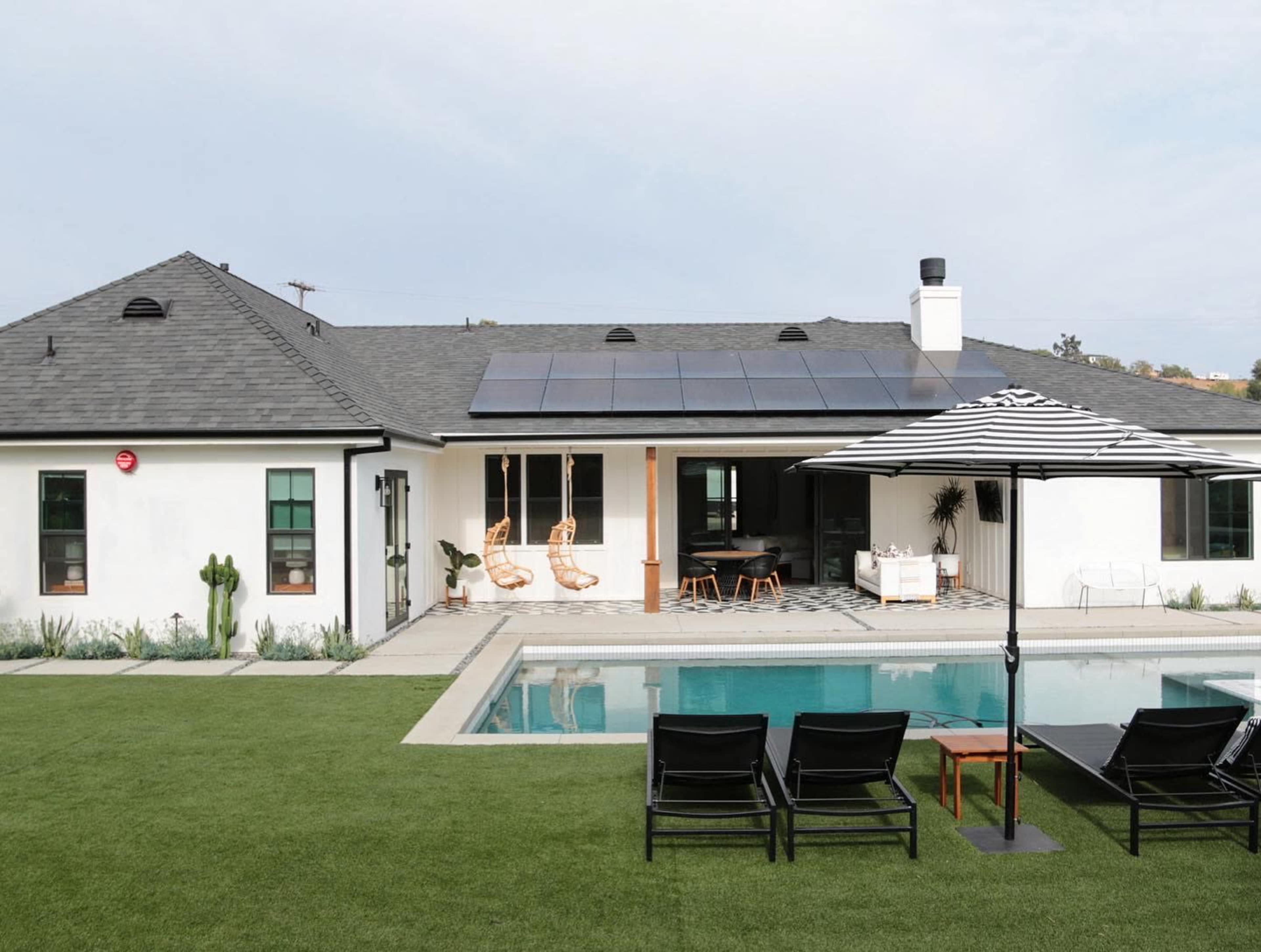 A modern house with solar panels features a swimming pool, lounge chairs, and a patio area surrounded by landscaped grass.