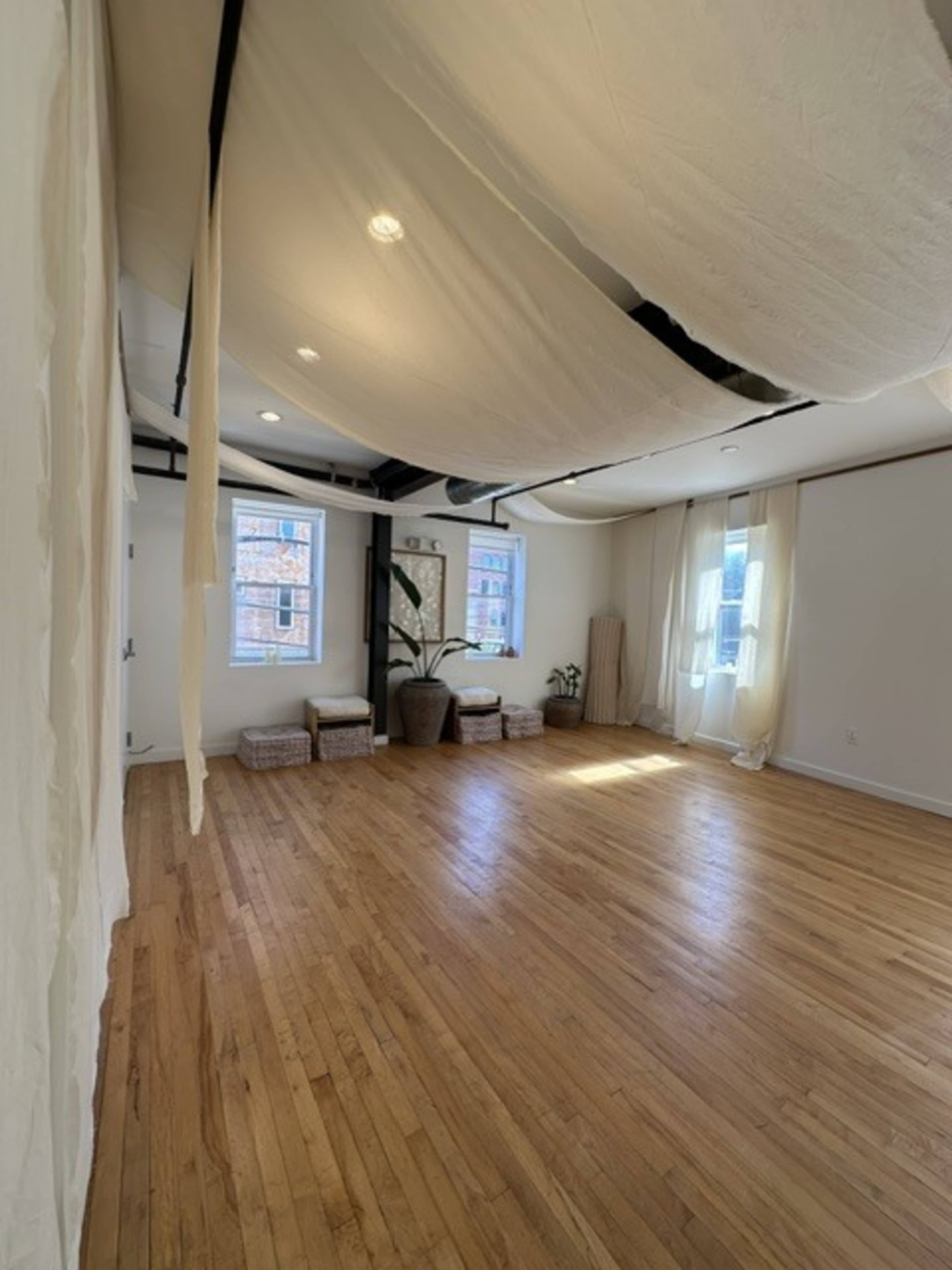 The image shows a spacious, well-lit room with wooden flooring, adorned with soft, draped fabric hanging from the ceiling and featuring a few decorative plants and wicker seating.
