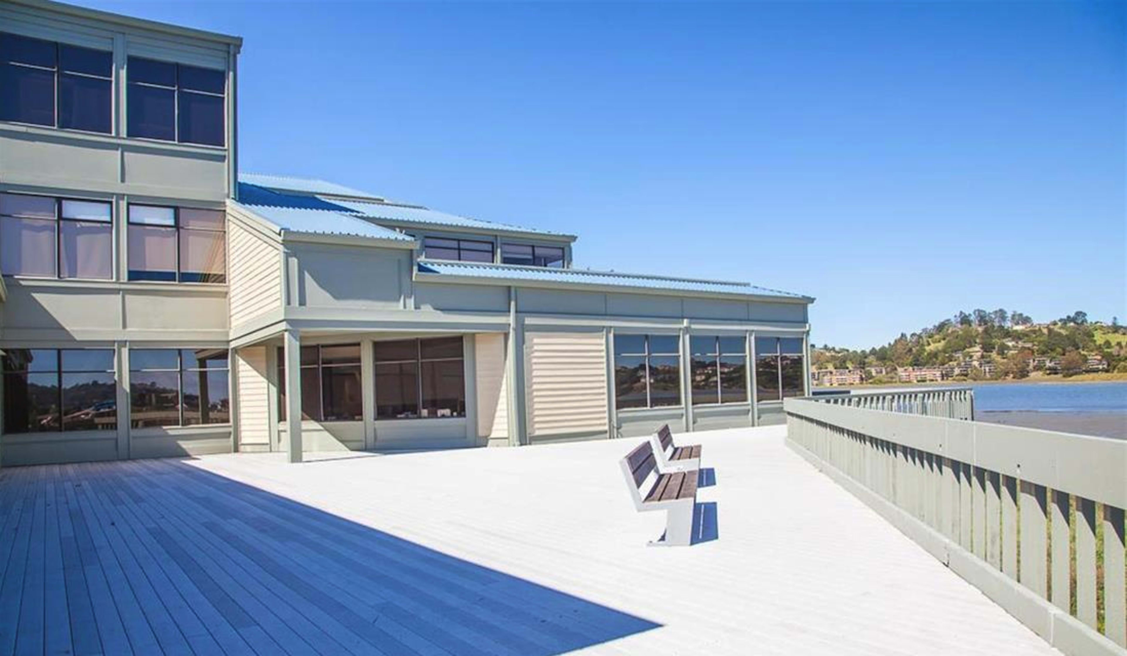 A modern building with large windows and a wooden deck extending toward a waterfront view.