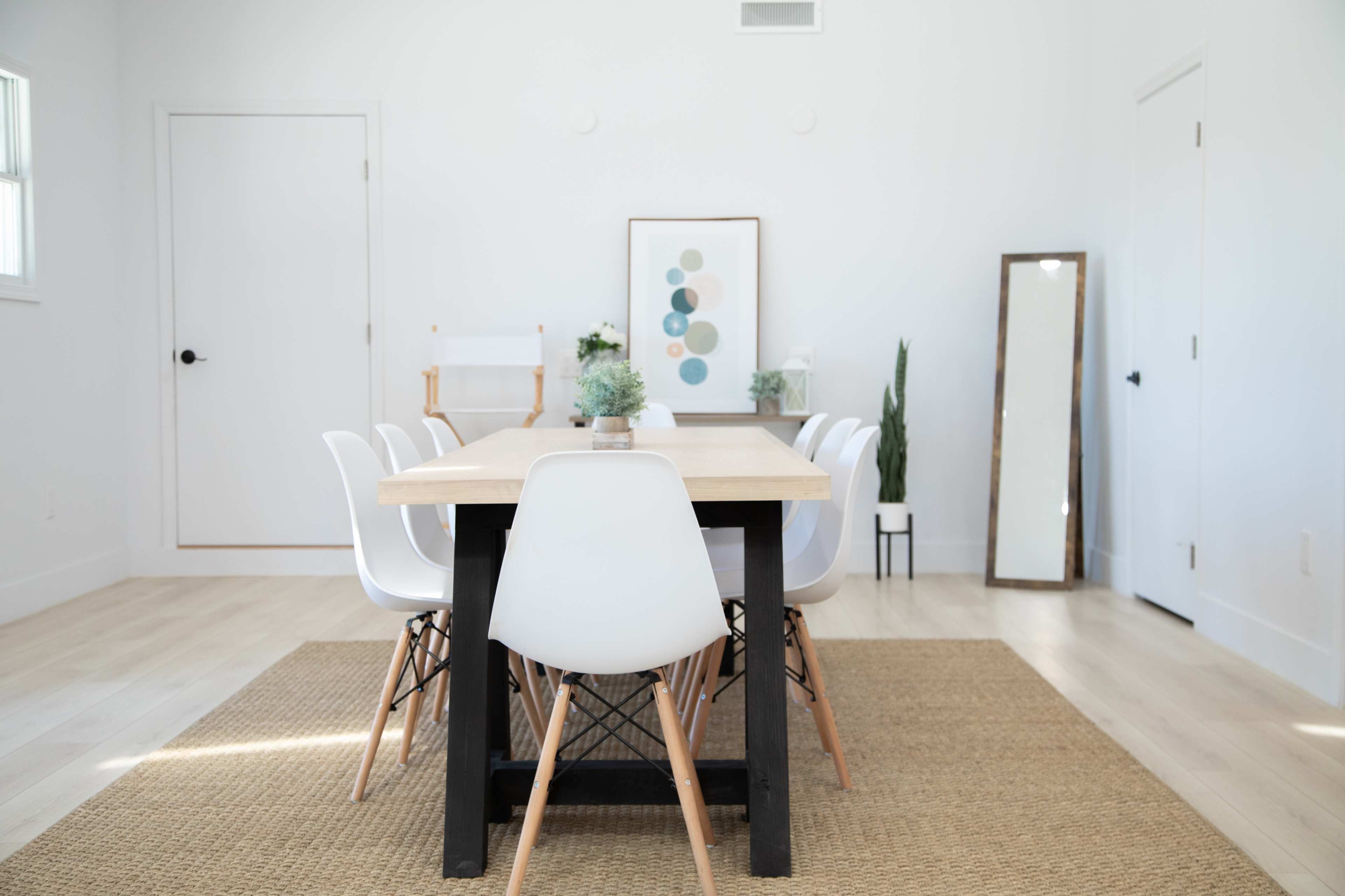 A light-filled dining area features a wooden table surrounded by white chairs, with a large mirror and decorative plants enhancing the minimalist decor.
